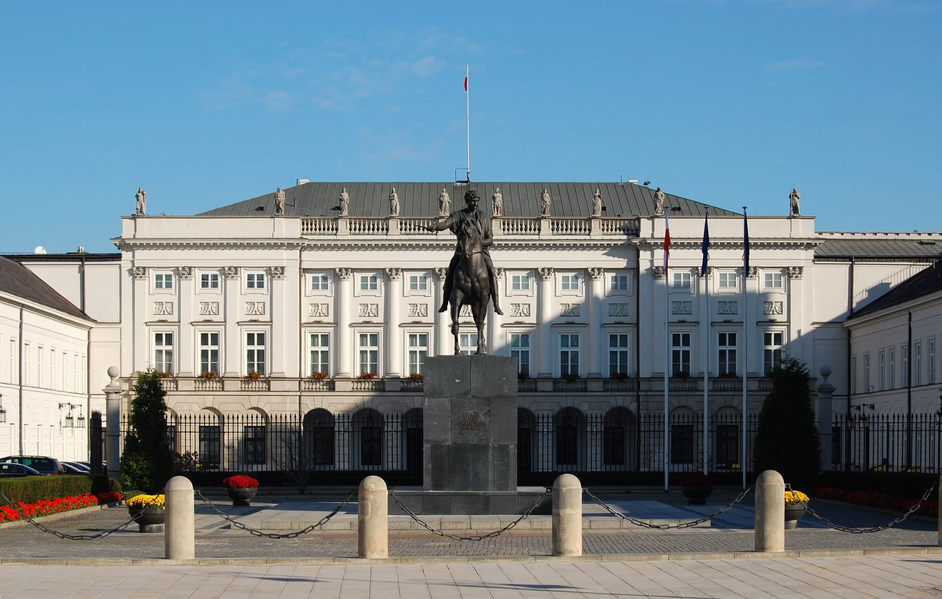 Kliknij, aby powiększyć. Na zdjęciu przedstawiono pałac. Posiada skrzydła i trzy kondygnacje. Przed pałacem znajduje się plac, na którym stoi rzeźba jeźdźcy na koniu. Przy pałacu biegnie chodnik.
