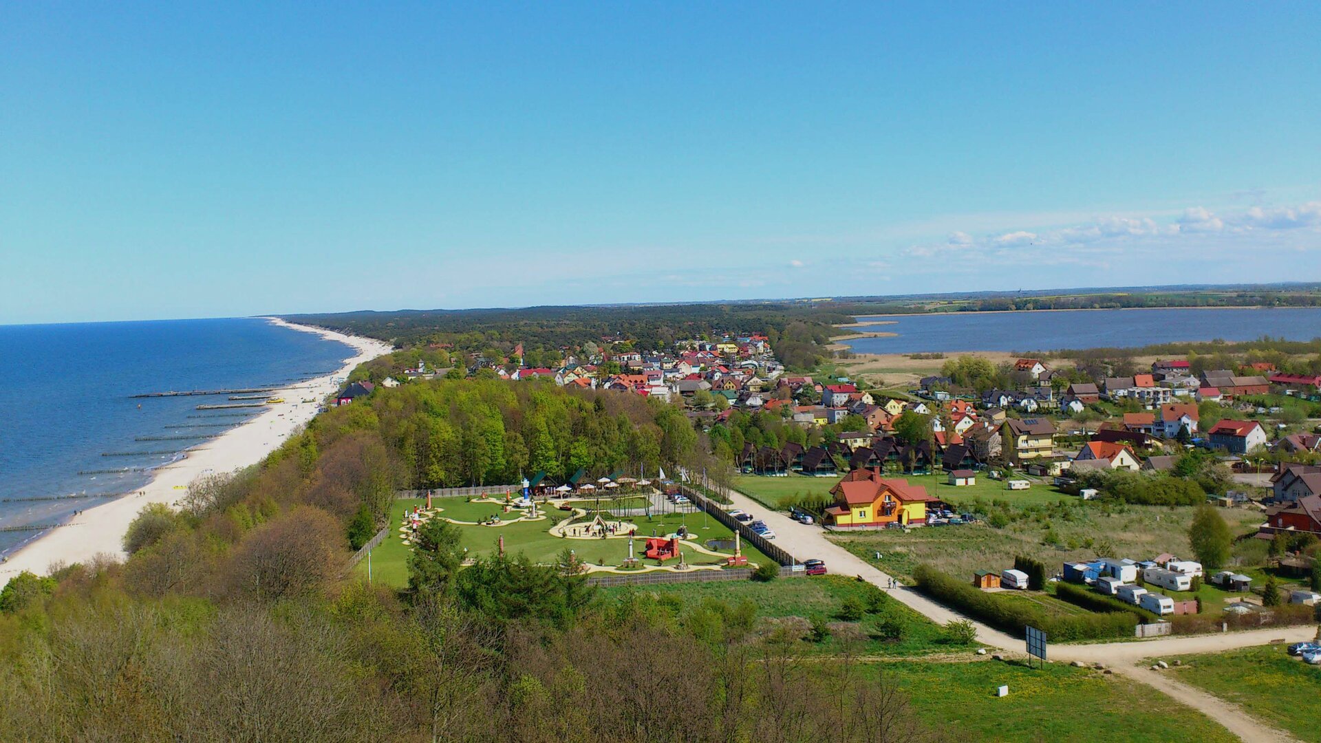 Na zdjęciu widok z góry na brzeg morski, plażę, teren zabudowany i tereny zielone oraz jezioro przybrzeżne w niewielkiej odległości od morza.