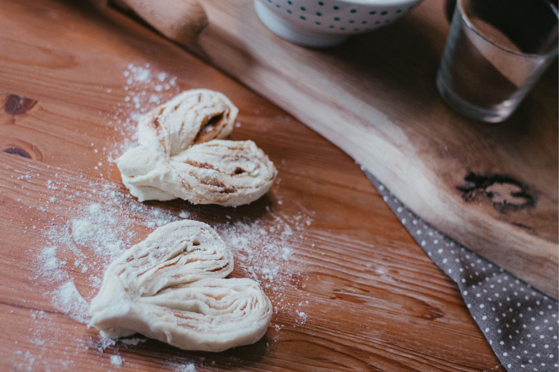 Zdjęcie przedstawia kontynuację kroku szóstego formowania serduszek. Ujęcie wykonane jest od góry pod skosem. Na pierwszym planie w lewej części grafiki widoczne są dwa przecięte i rozchylone na boki rulony ciasta. Uformowane są one na kształt serca i oprószone mąką. Widoczne w przekroju są warstwy ciasta, a pomiędzy nimi znajdujący się brązowy cukier cynamonowy. Całość ułożona jest na drewnianym ciemnobrązowym blacie. Dookoła rozsypana jest biała mąka. W lewej górnej części zdjęcia widoczna jest jasnobrązowa drewniana deska na której stoi biała miseczka w małe niebieskie kropki oraz przezroczysta szklanka wypełniona brązowym cukrem cynamonowym. W prawym dolnym rogu zdjęcia widoczna jest ciemna ścierka w białe małe kropki na której leży drewniana deska.