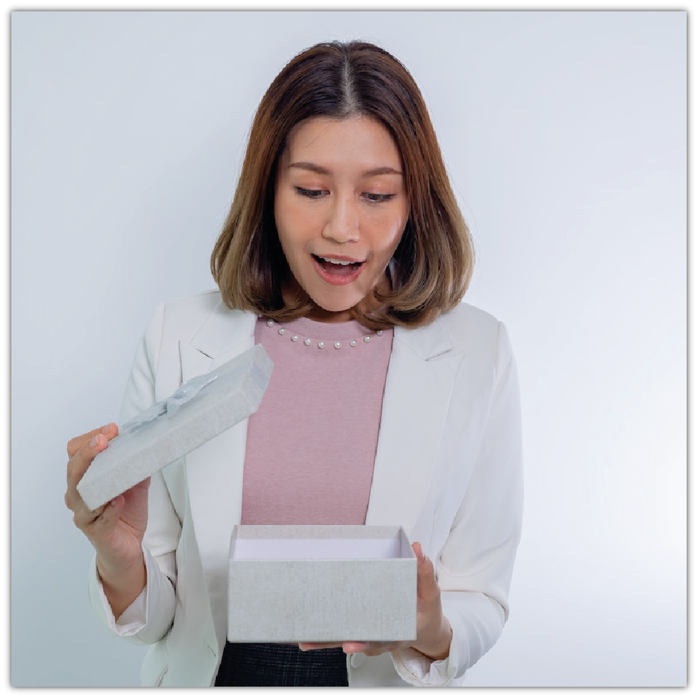 Zdjęcie przedstawia zaskoczoną, młodą kobietę, która otwiera białe pudełko prezentowe.