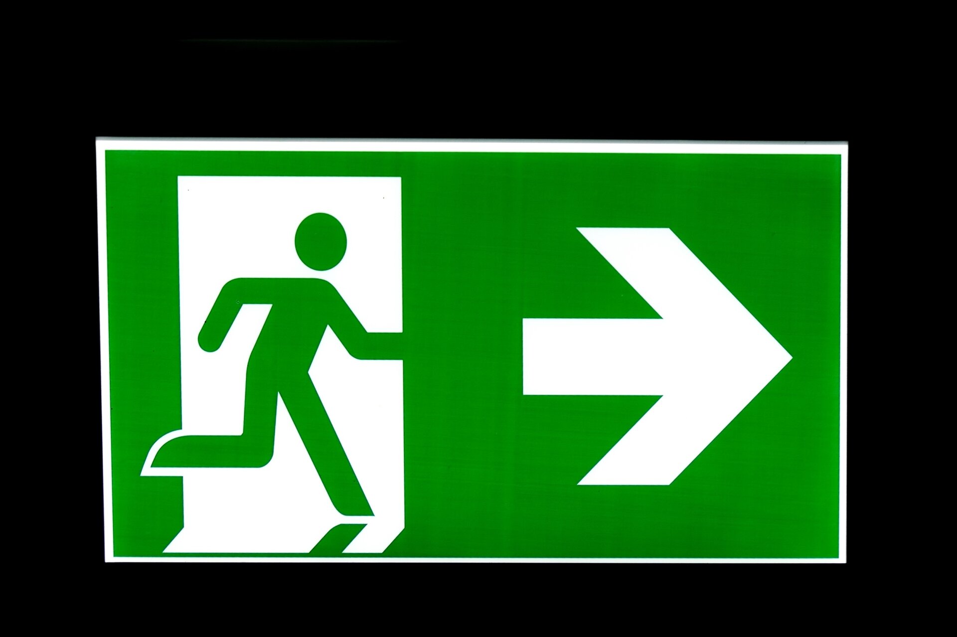 Zdjęcie przedstawia tabliczkę symbolizującą wyjście ewakuacyjne. Jest to zielona prostokątna tabliczka z białą obwódką. Po lewej stronie jest piktogram zielonego człowieka wybiegającego przez białe drzwi. Po prawej stronie znajduje się biała strzałka w prawo.