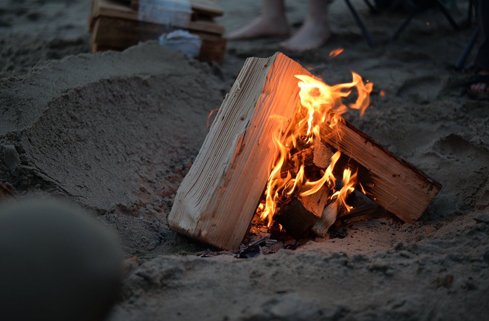 Zdjęcie przedstawia niewielkie zagłębienie w piasku, w którym pali się małe ognisko. Widoczne są jasne polana drewna obejmowane przez żółto‑pomarańczowe płomienie. W tle widoczne są stopy osoby siedzącej na rozkładanym krześle, przed którą leżą drewniane polana.