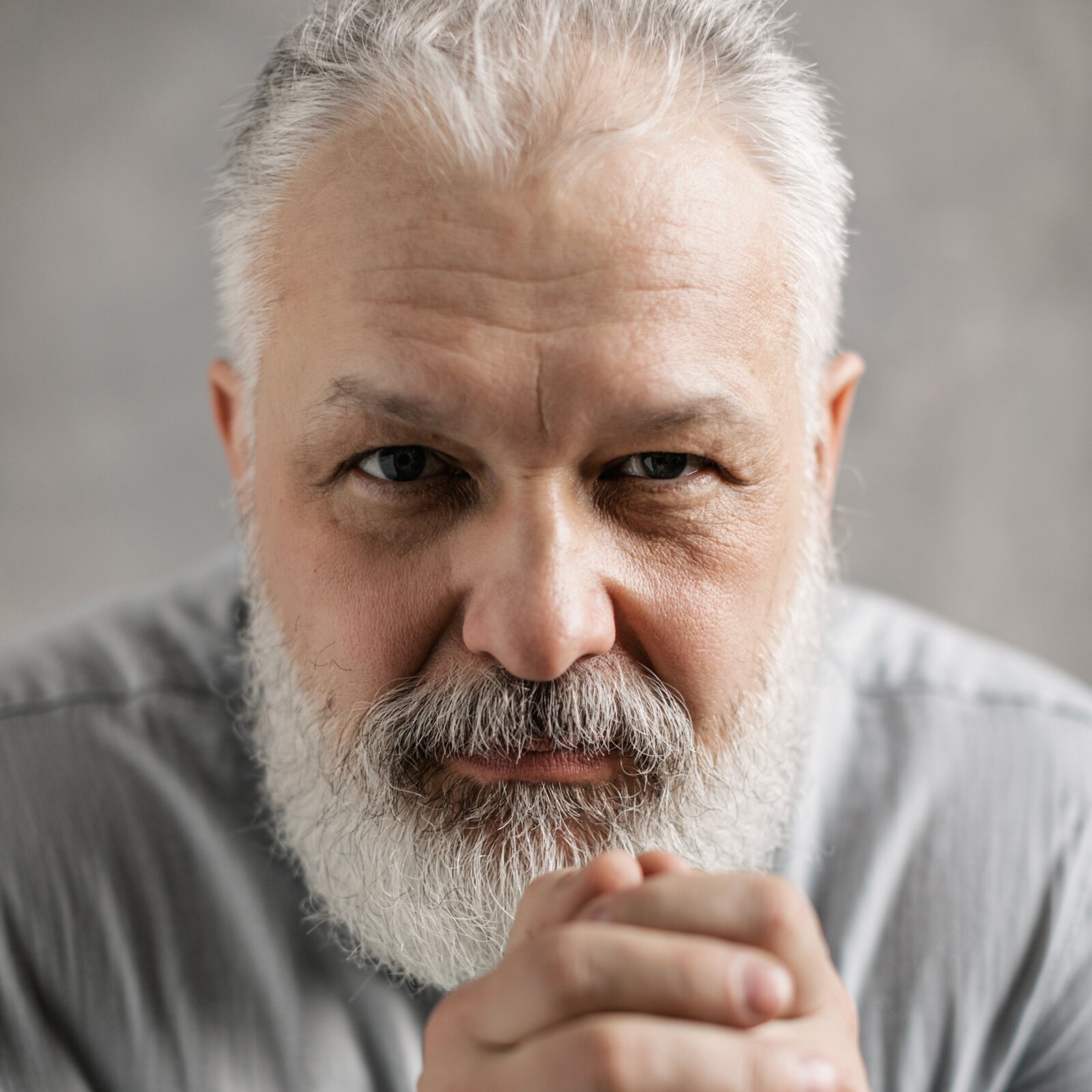 Zdjęcie przedstawia starszego, grubszego, siwego mężczyznę, który opiera się na ręce. Ma niebieskie oczy i spory nos. Ma zmarszczki na czole. Nosi brodę i wąsy.