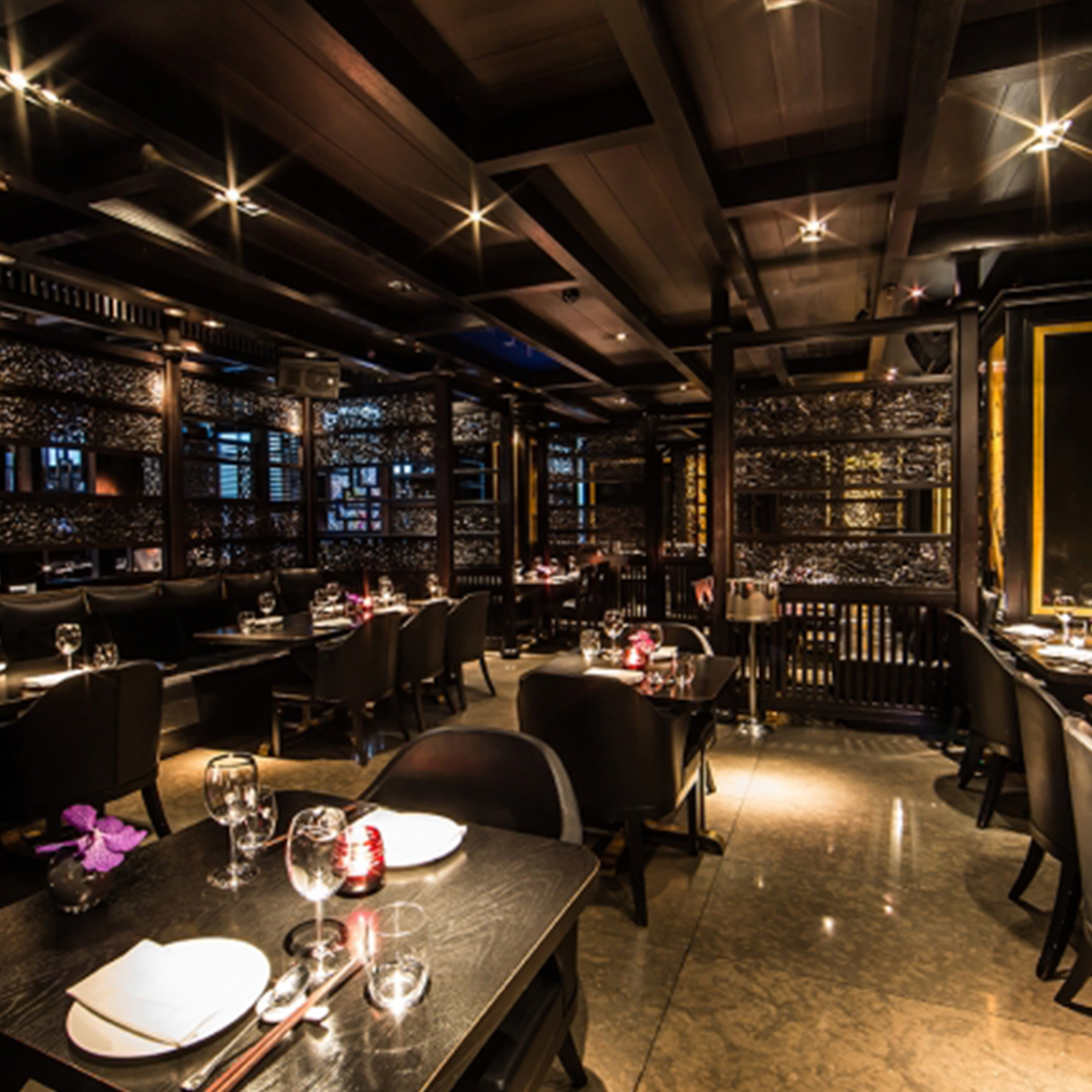Zdjęcie przedstawia wnętrze eleganckiej restauracji. Widoczne są stoliki z ciemnego drewna, przy których stoją brązowe fotele. Na stolikach ustawione są nakrycia, kieliszki, wazoniki z orchideą oraz palące się lampiony. Sala restauracyjna jest podzielona na strefy za pomocą ażurowych, drewnianych ścianek. W pomieszczeniu panuje półmrok.