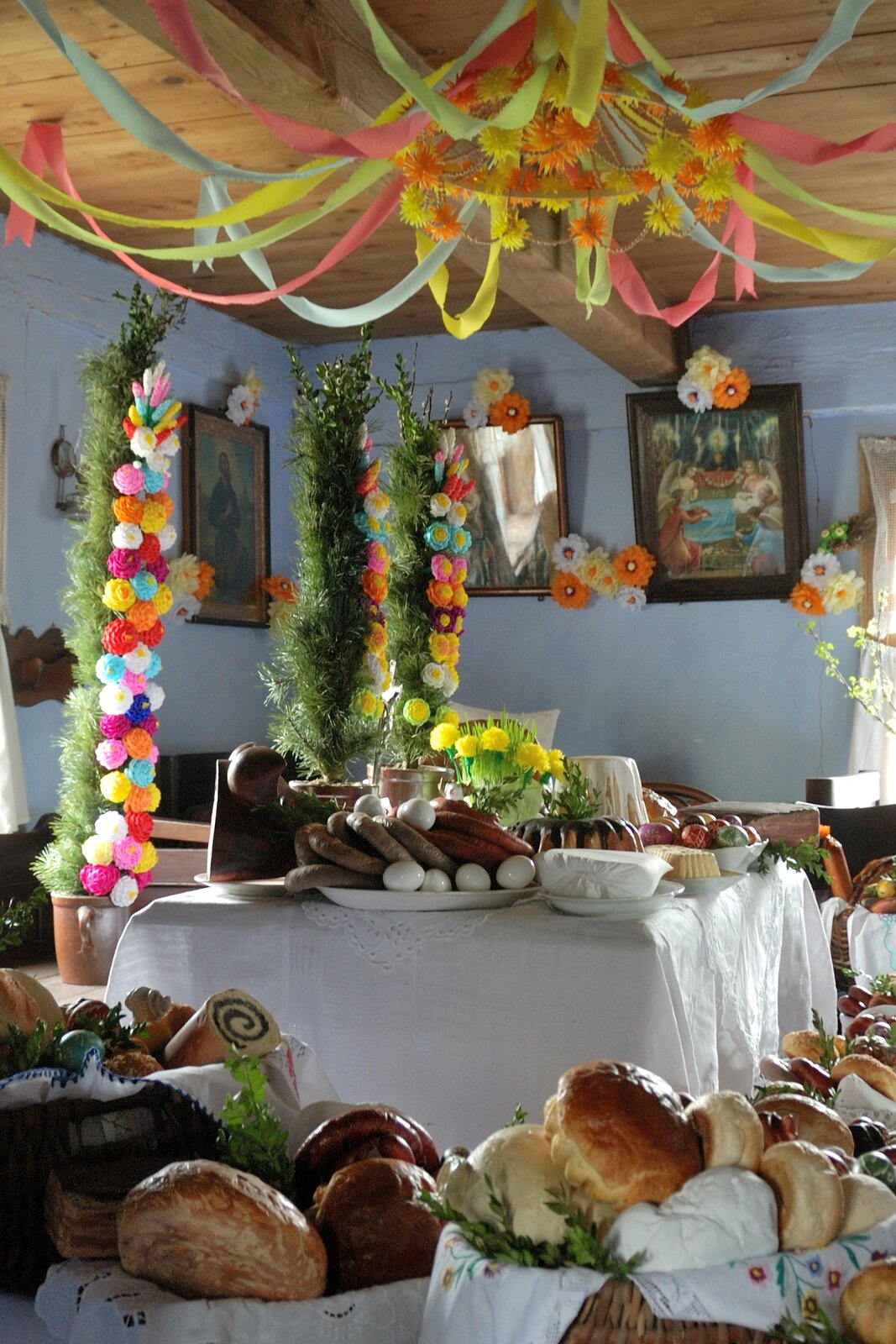 Zdjęcie przedstawia pomieszczenie, w którym ustawiono tradycyjne wielkanocne potrawy oraz dekoracje. Na środku znajduje się mały, kwadratowy stół z białym obrusem. Na nim leżą talerze z ułożonymi potrawami. Są to kiełbasy, gotowane jajka, babka, kolorowe pisanki oraz wysokie kolorowe palmy wielkanocne. Nad stołem, pod sufitem wisi okrągła dekoracja z żółtymi i pomarańczowymi kwiatami i paskami z bibuły. Dookoła stołu ustawiono ekspozycję wypieków - chlebów, bułek i słodkości, w drewnianych koszyczkach. Na ścianach pomieszczenia powieszono kilka obrazów z wizerunkami świętych.