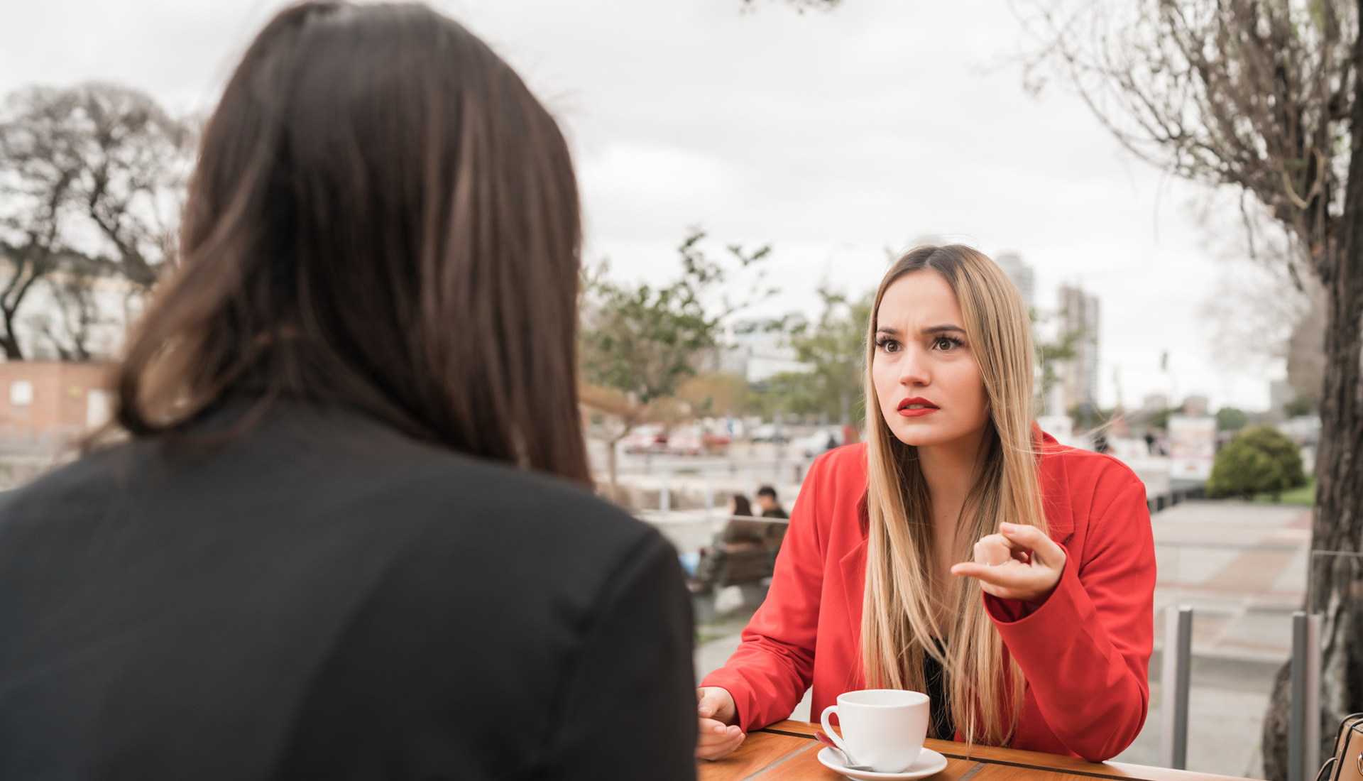 Zdjęcie przedstawia dwie młode kobiety, siedzące przy stoliku w ogródku kawiarni. Po prawej stronie widoczna jest kobieta, zwrócona lekko w lewo, patrząca na dziewczynę siedzącą na przeciw. Ma poirytowaną minę, długie, rozpuszczone blond włosy i jest ubrana w czerwony żakiet. Jej praw ręka leży na stoliku, zaś lewa, oparta łokciem o stolik, ma wysunięty palec wskazujący skierowany na osobę siedzącą naprzeciw. Przed nią, na blacie, stoi biała filiżanka na spodku. Po lewej stronie widać tył drugiej dziewczyny, z dłuższymi, rozpuszczonymi ciemnymi włosami, ubranej w czarny żakiet. W tle widoczny jest rozmazany chodnik, budynki i drzewa. 