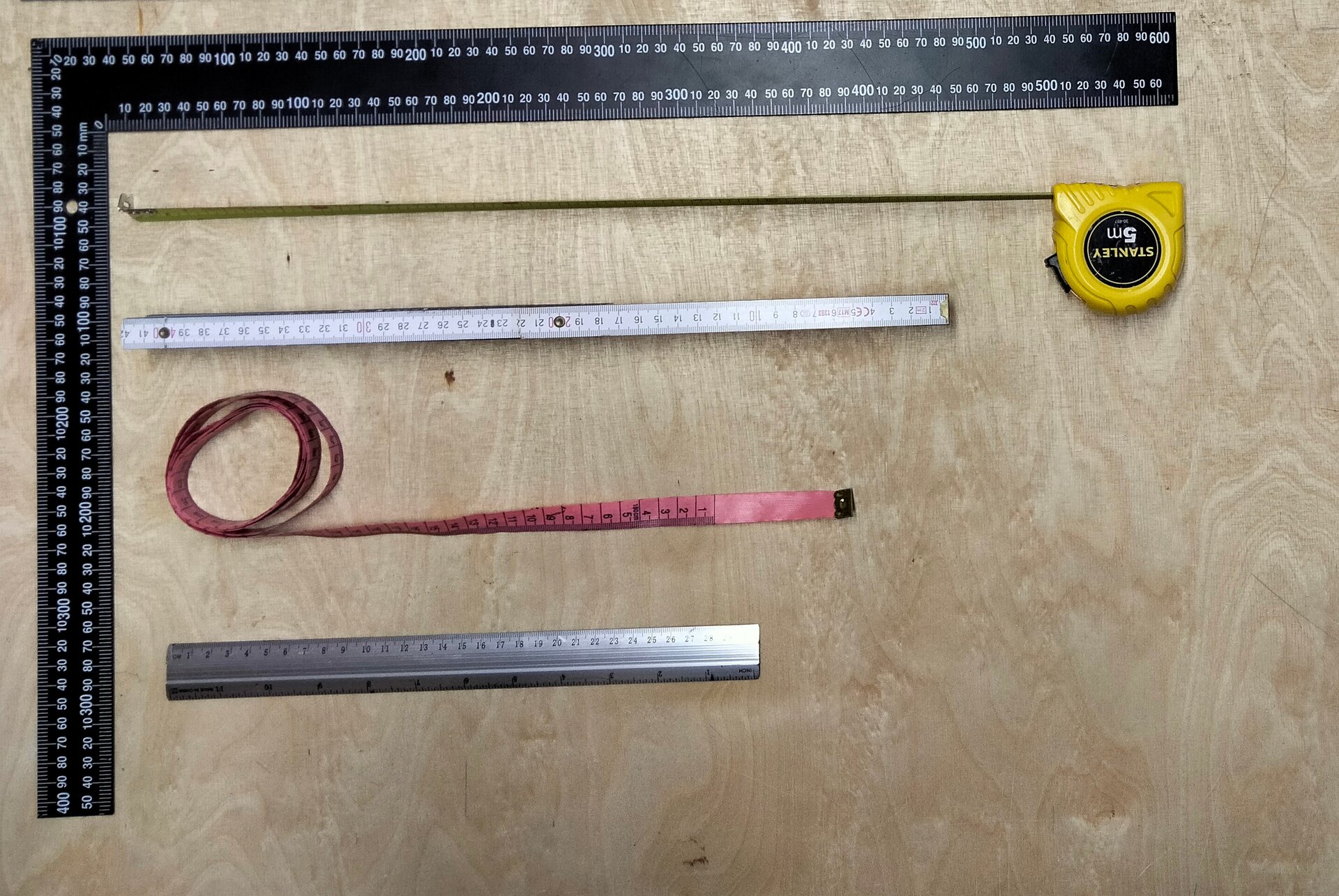 Na zdjęciu przedstawiono pięć różnych przyborów pomiarowych, czyli: kątownik, przymiar taśmowy zwijany, przymiar stolarski składany, przymiar krawiecki, linijkę. Przybory te są umieszczone równolegle do siebie.