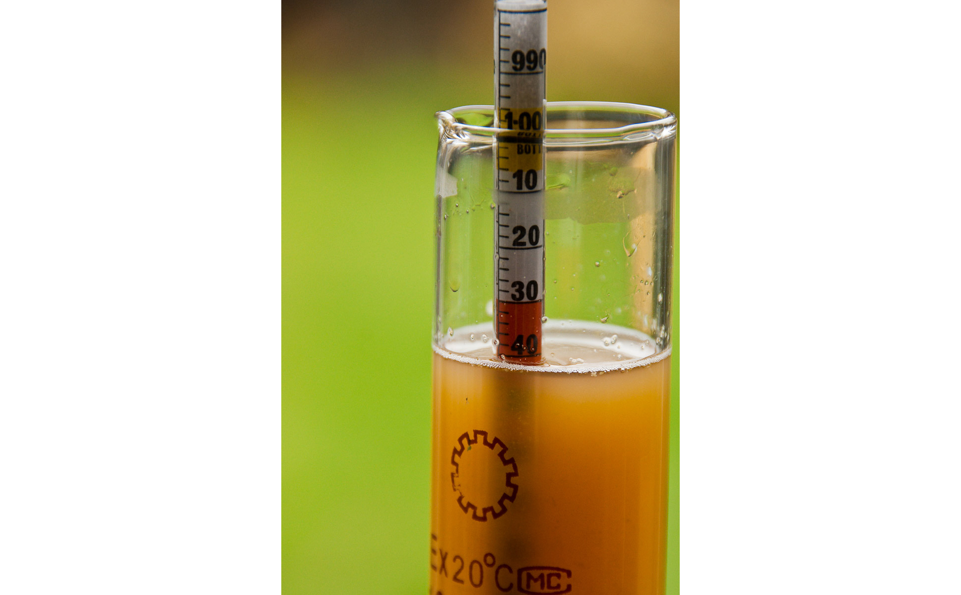Zdjęcie przedstawia areometr włożony do szklanej zlewki z cieczą. Tło zielone, rozmyte. Ciecz brunatna, nieklarowna. Na areometrze widoczna skala. Od góry liczby: 990, druga niewidoczna, zakryta częściowo przez brzeg zlewki, następnie: 10, 20, 30, 40. Powierzchnia cieczy wskazuje na liczbę 40.