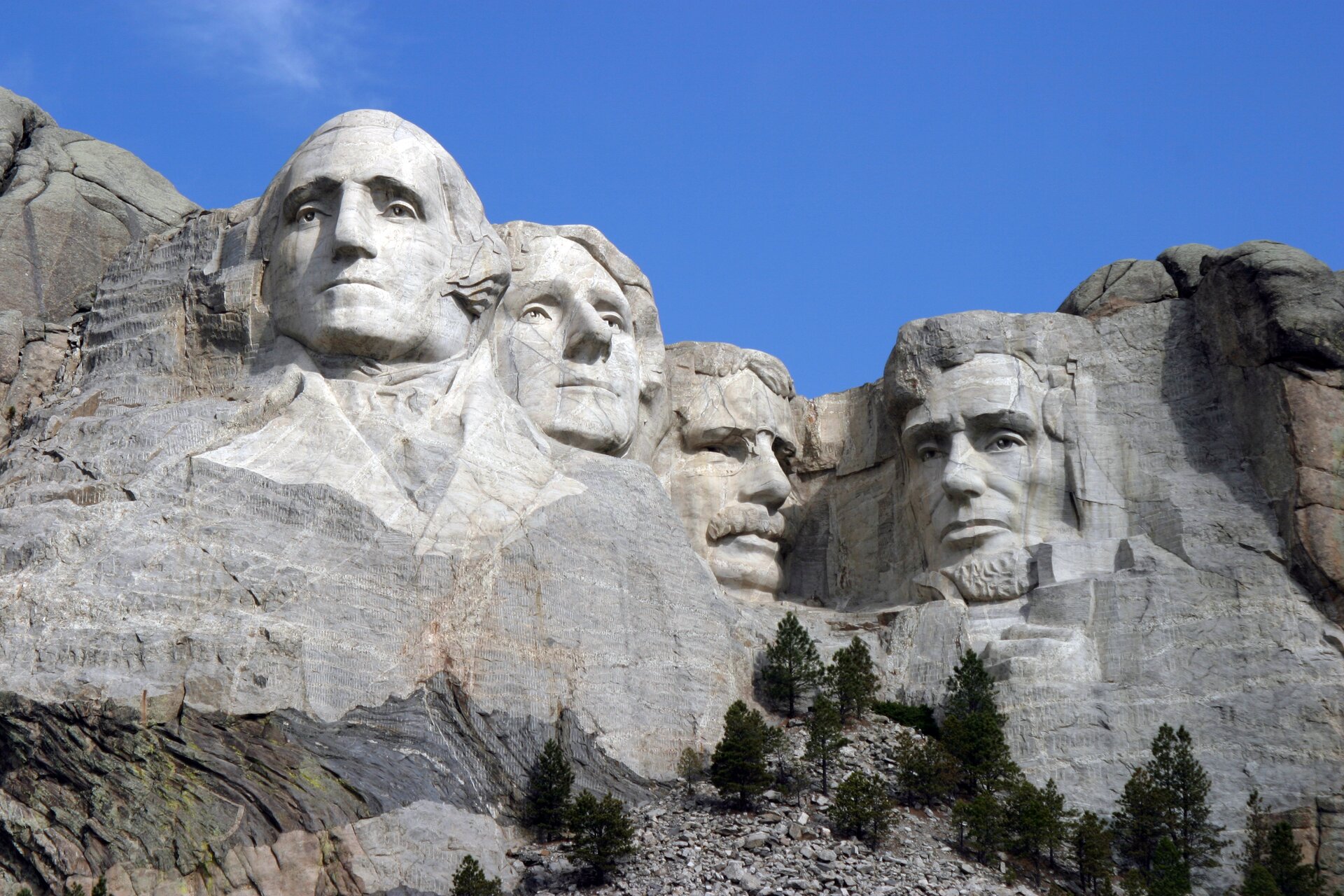 Ilustracja przedstawia „Pomnik Mount Rushmore”, autorstwa Gutzona Borglumy. Ukazuje wykute w skałach głowy czterech prezydentów USA. Od lewej są to: George Washington, Thomas Jefferson, Theodore Roosevelt oraz Abraham Lincoln. Mimika postaci wskazuje na powagę.
