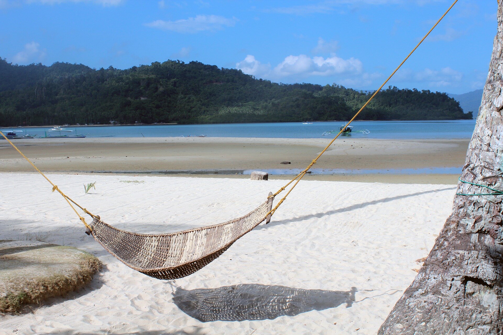 Fotografia ukazuje spokojną zatokę z piaszczystą plażą, na której wisi hamak rozpięty między drzewami. W tle spokojne morze i drzewa, które tworzą malowniczy krajobraz pełen spokoju i harmonii.