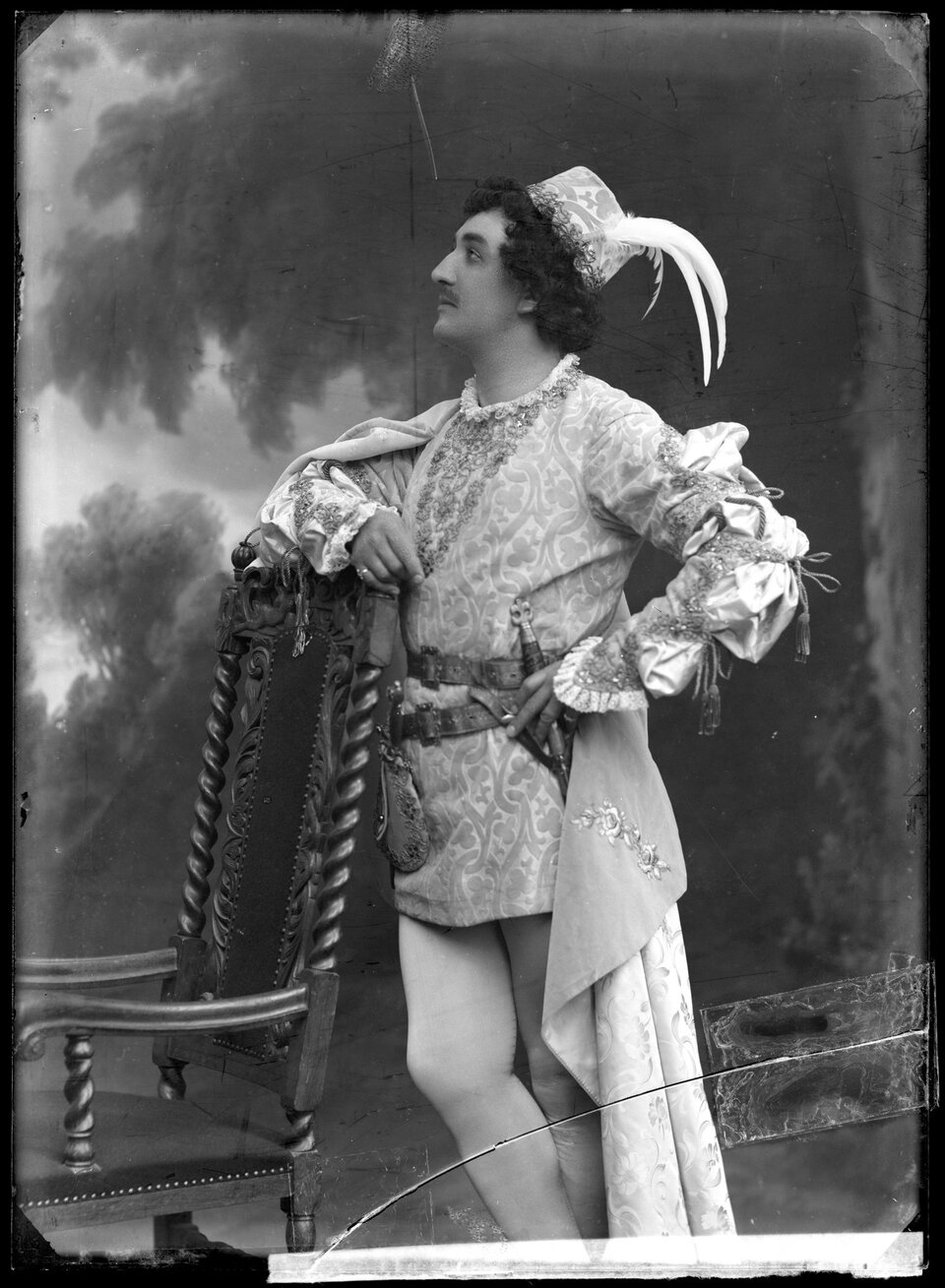 Czarno‑białe zdjęcie przedstawia młodego człowieka stojącego bokiem, opartego prawym ramieniem o rzeźbione krzesło. Mężczyzna jest ubrany w ozdobny kaftan z bufiastymi, wiązanymi rękawami, przy boku ma sztylet umocowany na skórzanym pasie. Nosi białe rajtuzy. Z ramienia zwisa peleryna. Na głowie ma niewielki kapelusik z białymi piórami. Mężczyzna patrzy przed siebie, ma kręcone włosy sięgające do szyi, niewielki wąsik. Twarz widać z profilu. W tle zamazane drzewo.