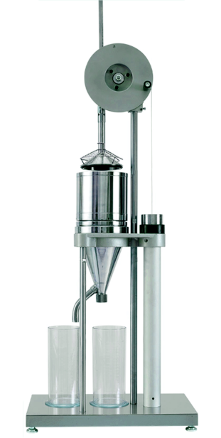 Zdjęcie ukazujące urządzenie do oznaczania stopnia zmielenia metodą szopera riglera. Na prostokątnej podstawie urządzenia stoją dwie przezroczyste zlewki na próbki i podłużna noga, na szczycie której zamontowano metalowy lejek, tubę i pracę do toczenia próbek. Nad konstrukcją znajduje się przymocowana do niej dwoma wspornikami, obrotowa konstrukcja wprawiająca tłok w ruch.