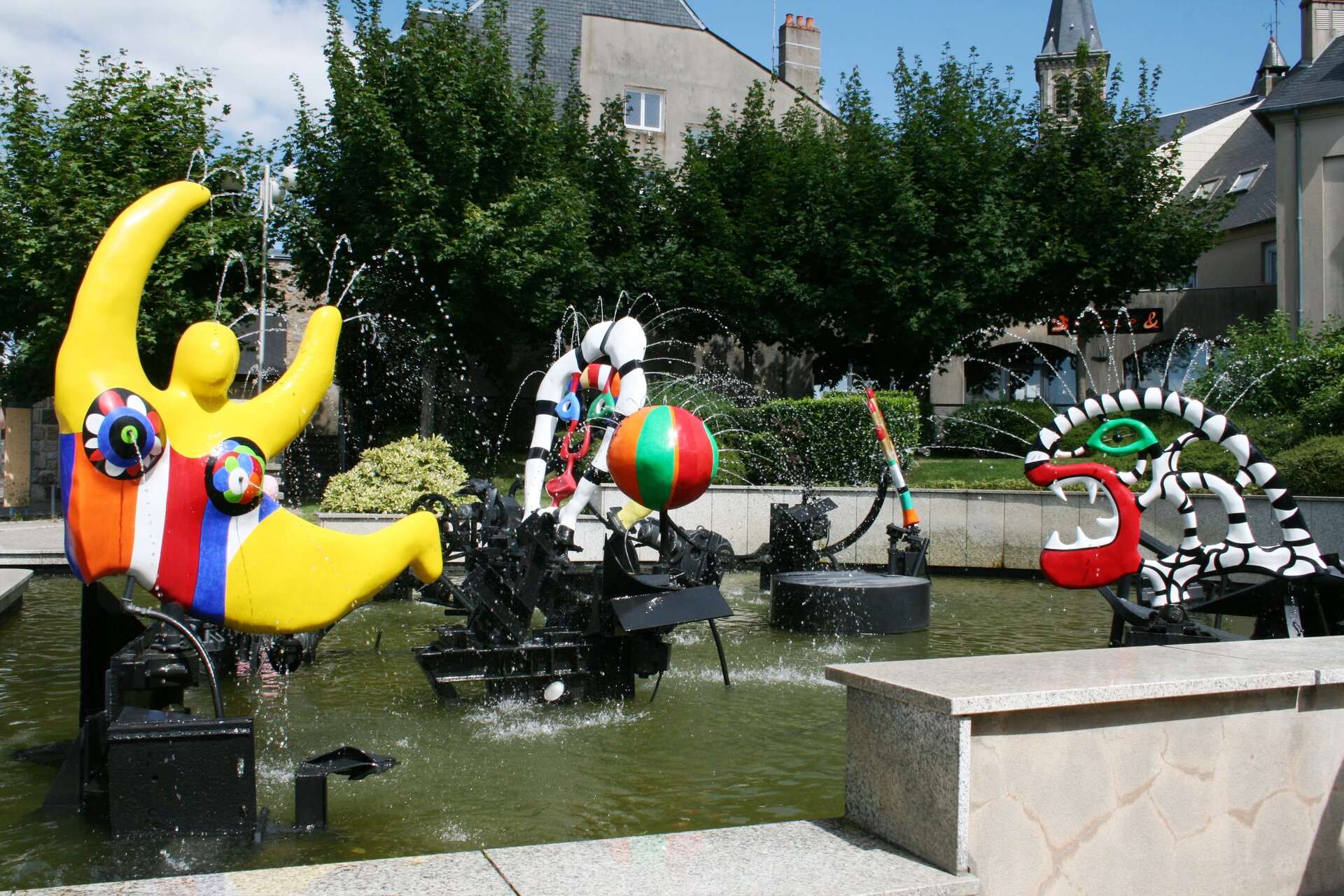 Fotografia przedstawia widok na fontannę na tle domów oraz drzew o zielonych liściach. Mur otaczający fontannę jest jasnoszary, pokryty od góry płytkami. W fontannie widoczne są kolorowe rzeźby stojące na czarnych podestach. Pierwszy plan stanowi żółta rzeźba, jej kształt przypomina postać szykującą się do skoku uniesionymi do góry rękami oraz złączonymi nogami. Figura ma na sobie kolorowy strój w pomarańczowe, białe, czerwone i niebieskie pasy. Z piersi postaci tryska woda. Z prawej strony stoi rzeźba przypominająca dinozaura o czerwonych ustach z której wystają ostre szpiczaste zęby. Ma zielone oczy oraz białą konstrukcję w czarne prążki. Drugi plan stanowi rzeźba przedstawiająca piłkę plażową w zielono‑pomarańczowo‑czerwone pasy. Za nią, z prawej strony - kolejna rzeźba swoim kształtem przypomina spłaszczone koło ratunkowe i jest biała w czarne paski. Wewnątrz niej umieszczono elementy przypominające oczy, nos i usta.