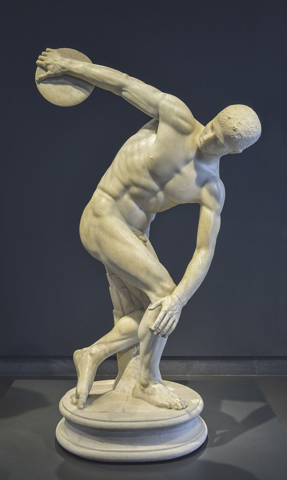 Zdjęcie przedstawia marmurową rzeźbę nagiego, atletycznie zbudowanego mężczyzny. Mężczyzna jest lekko pochylony do przodu, w prawej dłoni trzyma dysk, który zamierza rzucić przed siebie.