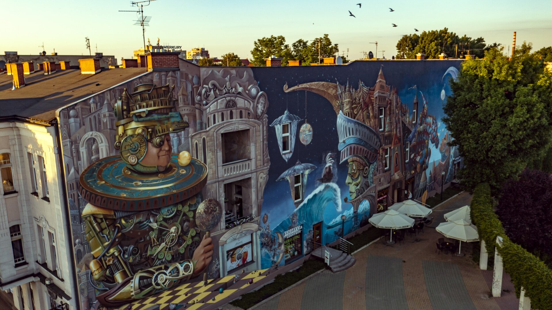 Ilustracja w formie prostokąta przedstawia fragment budynku, na którym namalowany jest mural. Przy ścianie budynku ustawione są cztery białe parasole, a pod nimi czarne stoły oraz krzesła. Obok nich znajdują się liczne drzewa. Mural wykonany jest przeważająco w odcieniach niebieskiego, brązowego oraz szarego. Ukazuje on abstrakcyjne postaci. Jedna z nich jest w połowie robotem, a w połowie człowiekiem. Posiada ludzką twarz oraz dłoń. Na jej głowie znajduje się złoty budynek oraz złoty mechanizm. Jedno oko mężczyzny zastąpione zostało złotą kulą. Na miejscu ucha znajdują się zegary. Pod szyją postaci zaprezentowana jest makieta układu słonecznego. Tułów mężczyzny składa się z wielu połączonych ze sobą mechanizmów, kabli, sprężyn czy rurek. W dłoni postać trzyma coś na wzór berła. Ręką mężczyzna opiera się na brązowo‑żółtej szachownic, na której rozsypane są małe, kolorowe kulki. Postać przedstawiona została na tle dużego, szarego budynku, który posiada wiele kolumn, fasad czy ornamentów. Po drugiej stronie muralu ukazane zostało “miasto” składające się z nachodzących na siebie wielu budynków, które umieszczone są również na wzniesieniu, które tworzy niebiesko‑czerwony mur umiejscowiony na zielonej głowie. Kształt muru powoduje, że wygląda on również jak korona na głowie postaci. Postać patrzy się na wprost, a za tyłem jego głowy wzrastają budynki. Przed budowlą znajduje się duża fala, na której płynie brązowo‑biały statek. Ta część muralu namalowana jest na niebieskim tle.