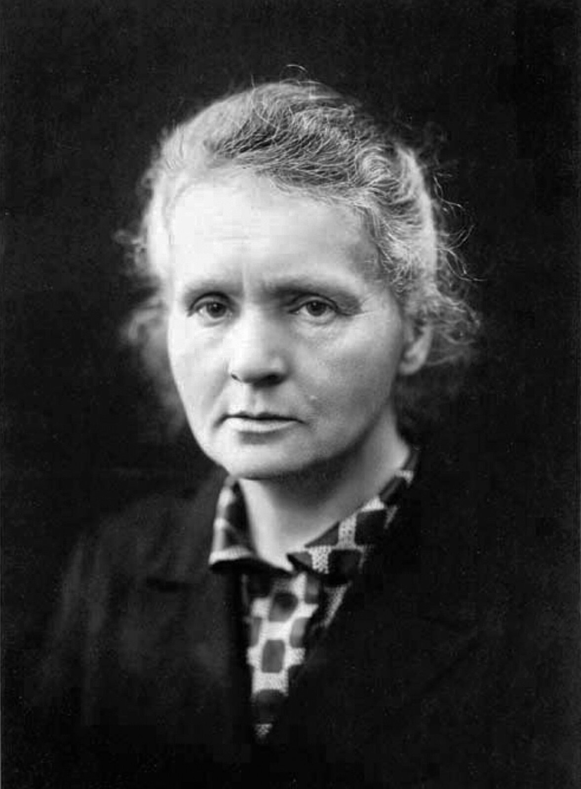Zdjęcie przedstawia Marię Skłodowską‑Curie. Jest to kobieta o siwych włosach upiętych w kok, ubrana w koszulę z kołnierzykiem w geometryczny wzór oraz czarną marynarkę. Ma poważny wyraz twarzy, patrzy przed siebie.