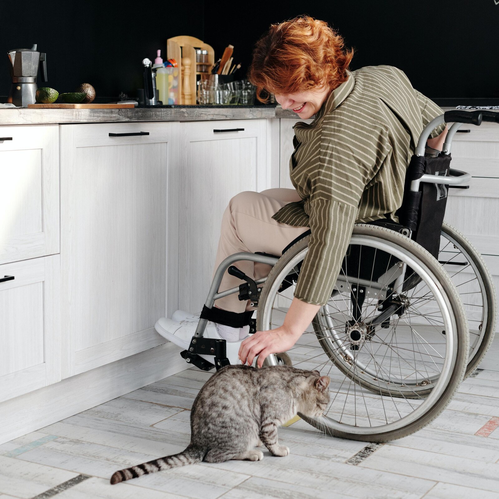 Ilustracja przedstawia uśmiechniętą osobę na wózku inwalidzkim, która pochyla się i głaszcze stojącego obok kota.