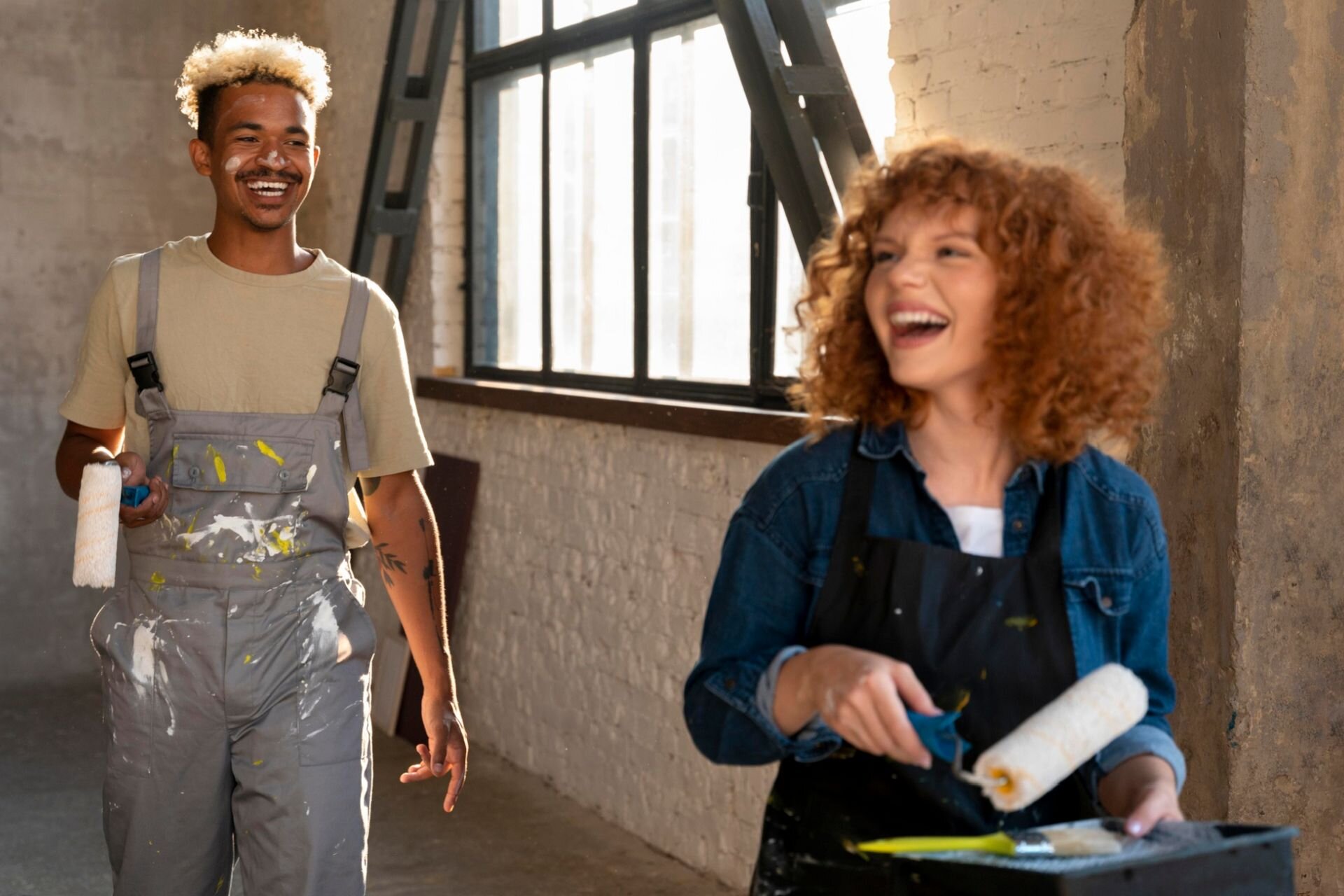 Ilustracja przestawia puste wnętrze na poddaszu w budynku z surowymi ścianami .Młoda uśmiechnięta kobieta i mężczyzna trzymają w rękach wałki malarskie.