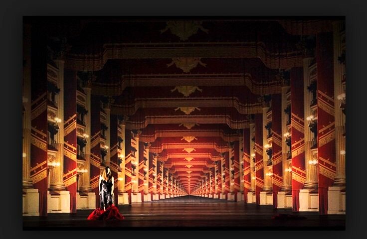 Zdjęcie przedstawia scenę operową, na której stoi kobieta. Za nią dekoracyjna kurtyna oraz kolumny po bokach ukazują nieskończenie długi korytarz. Dekoracja podporządkowana jest perspektywie zbieżnej, wszystkie elementy zmniejszają się aż do punktu zbiegu w oddali.