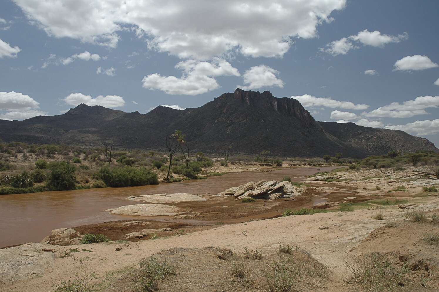 Fotografia. Na niej rzeka płynąca w poprzek fotografii. Po jednej stronie brzegu rzeki piasek, gdzieniegdzie kępy trawy, w oddali drzewa. Po drugiej stronie brzegu góry, u ich podnóża piasek z kępami traw. Rzeka ma kolor brązowy.  
