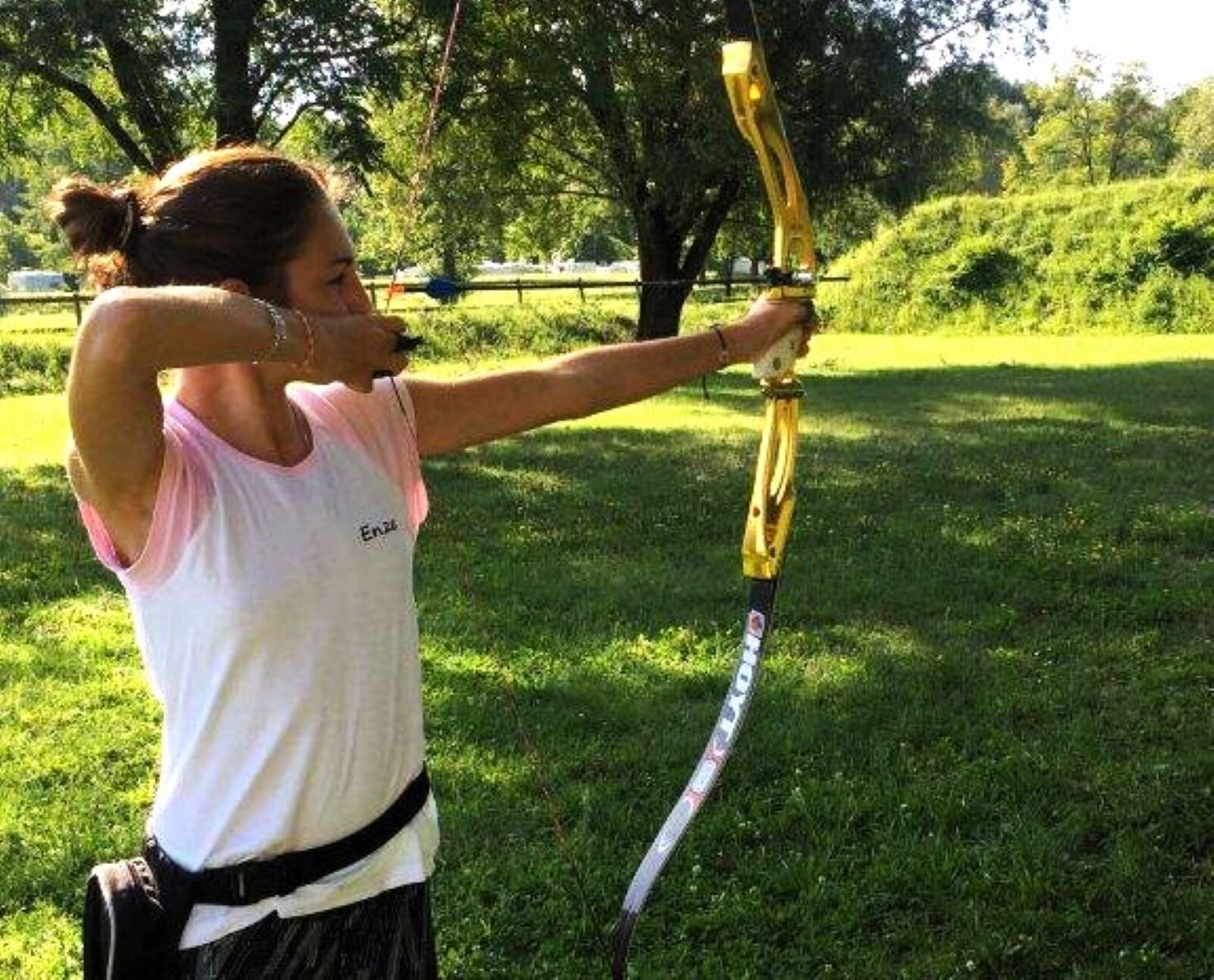 Zdjęcie przedstawia łuczniczkę w stroju sportowym, celującą z łuku. Jest słoneczna pogoda, dokoła zieleń. 