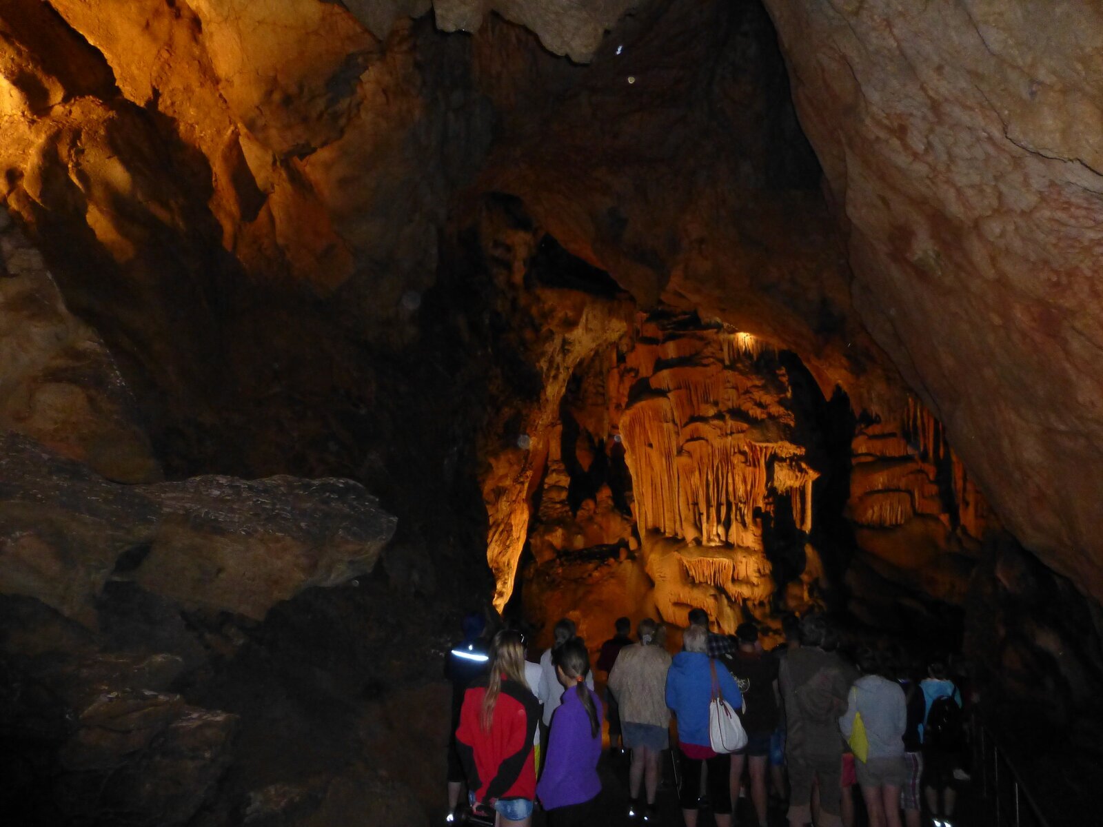Fotografia dużej, podświetlonej groty w jaskini z naciekami na ścianach. W jaskini znajduje się grupa zwiedzających ją turystów. 