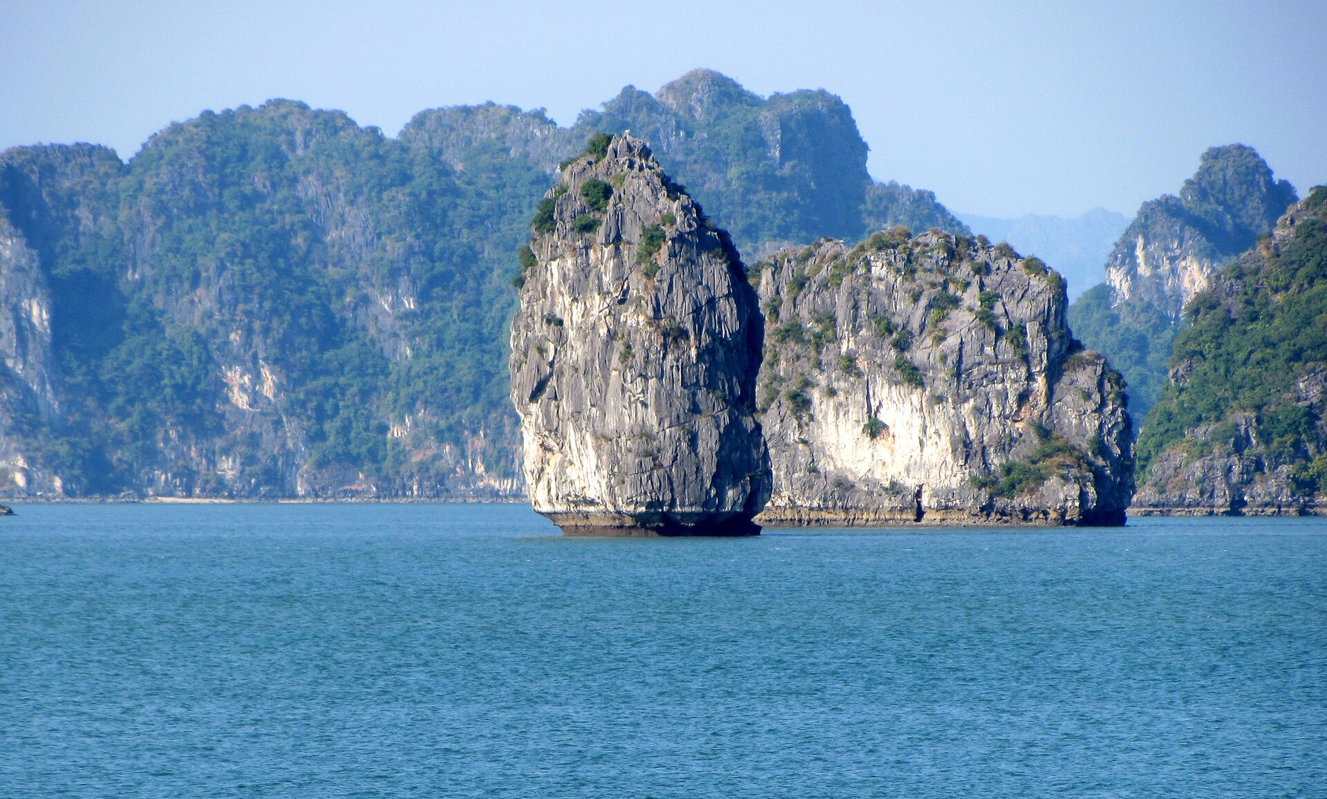  Zdjęcie przedstawia duży zbiornik wodny, na którego środku znajduje się skała o dużych rozmiarach. Swoim kształtem przypomina jajko. Za nią z wody wystaje skała niższa, ale dłuższa. W tle widać wysokie skały porośnięte drobną, zieloną roślinnością i niebieskie niebo.