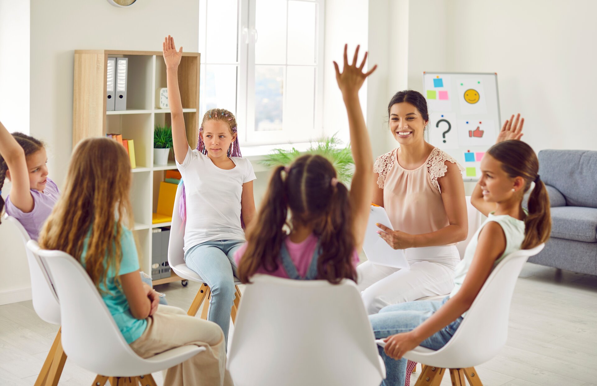 Zdjęcie przedstawia uczennice oraz nauczycielkę, siedzące w kręgu na białych krzesłach. Nauczycielka patrzy w stronę jednej z uczennic i uśmiecha się. Kilka uczennic podnosi rękę do góry. Zdjęcie wykonano w sali edukacyjnej.