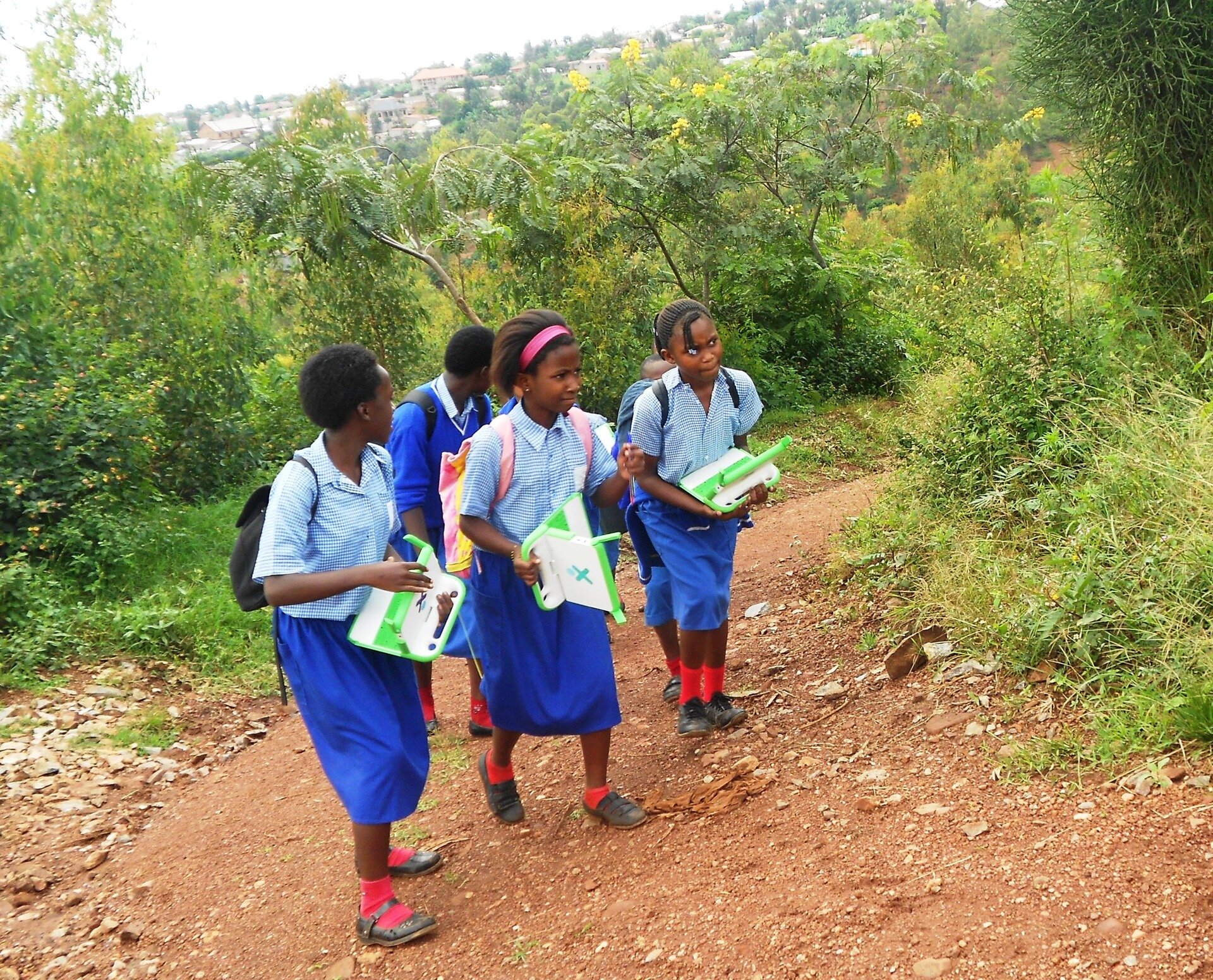 Na zdjęciu ciemnoskórzy uczniowie i uczennice w jednolitych strojach szkolnych. Bluzki i spódnice – dziewczęta, chłopcy noszą spodnie. Wszyscy mają w rękach laptopy. Dzieci idą drogą wśród zieleni.