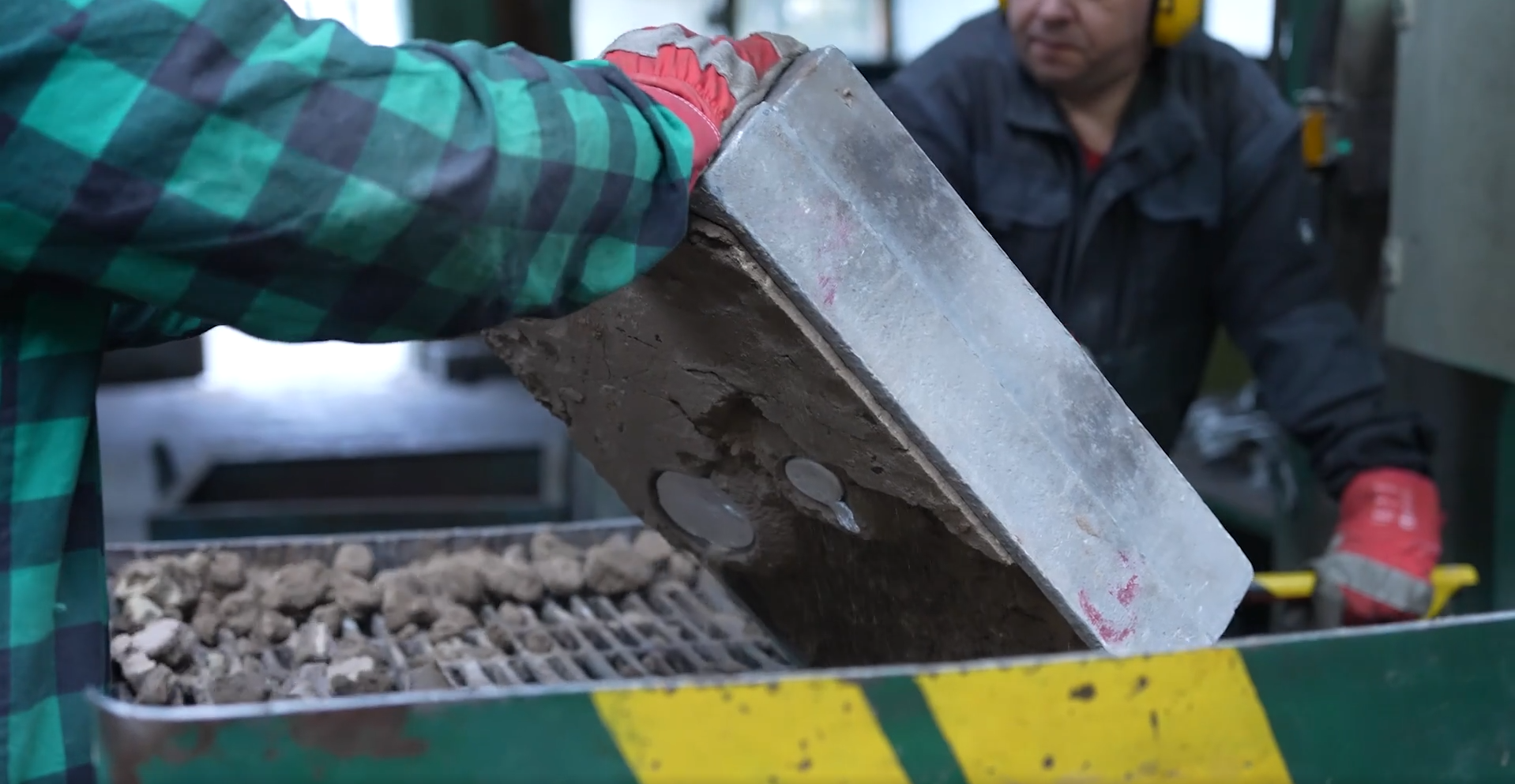 Na zdjęciu widoczne jest wybijanie odlewów na kratach wstrząsowych. W centralnej części zdjęcia znajduje się stalowa krata. Krata osadzona jest w prostopadłościennej obudowie. Przy obudowie stoi dwóch pracowników. Jeden z pracowników unosi dużą prostopadłościenną pokrywę. Na kracie widoczne są kawałki odlewów. Kawałki mają nieregularny kształt.