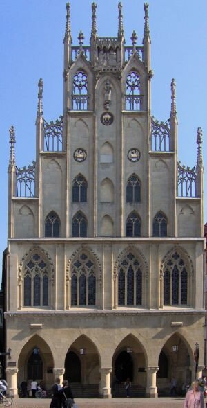 Zdjęcie przedstawia ratusz. Na parterze znajdują się łuki. W każdym łuku jest wisząca latarnia oraz drzwi. Okna wyżej są duże, każde posiada podział na trzy pionowe części a na górze mają otwory w kształcie koniczyn. Jeszcze wyżej są mniejsze okna oraz pionowe kolumny. Najdłuższa jest na środku. Im bliżej boków, tym niższe. Na górze każdej z nich są długie zakończenia. Przed budynkiem są ludzie na placu.