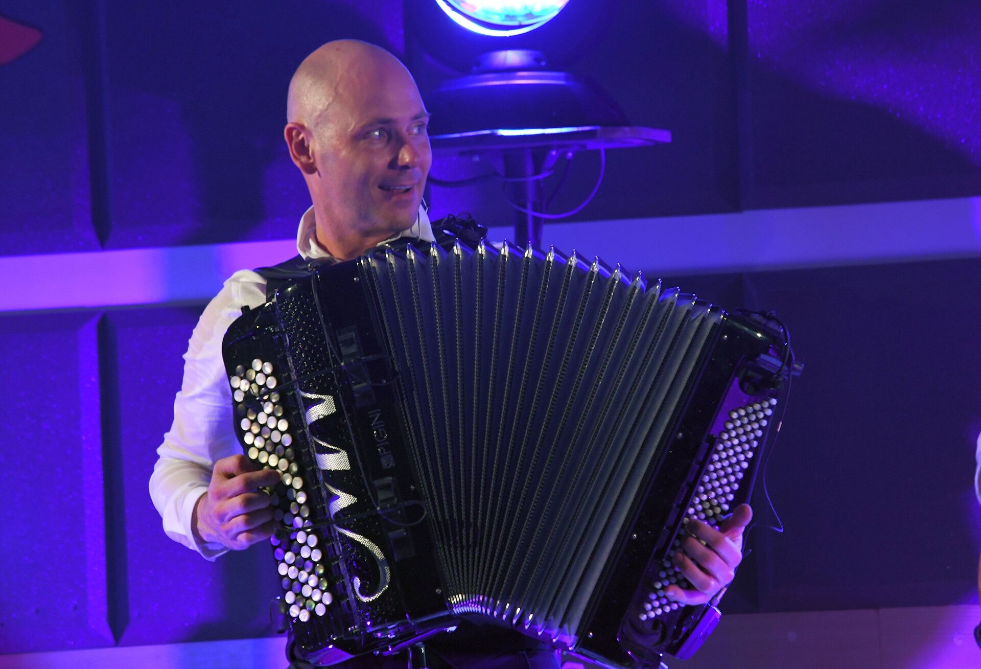 Na zdjęciu łysy, uśmiechnięty mężczyzna grający na akordeonie, który zasłania cały jego tułów. Widoczny jest od pasa w górę. Za nim jest ściana i lampa dająca niebieskie światło.