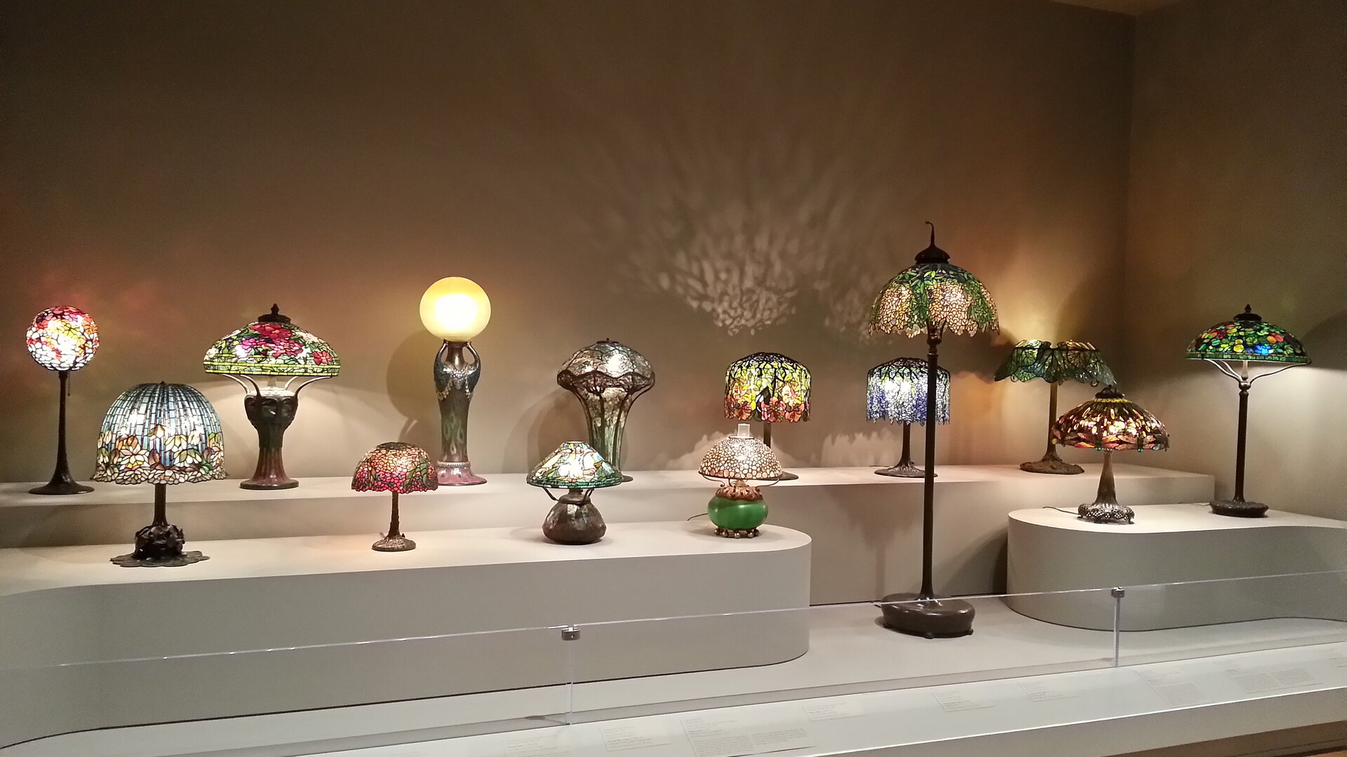 Zdjęcie przedstawia zbiór lampek nocnych, wykonanych techniką Tiffany'ego. Są ustawione na podestach na różnych poziomach. Wszystkie są włączone. Światło przenikające przez ich klosze tworzy efekty barwne na ścianie,