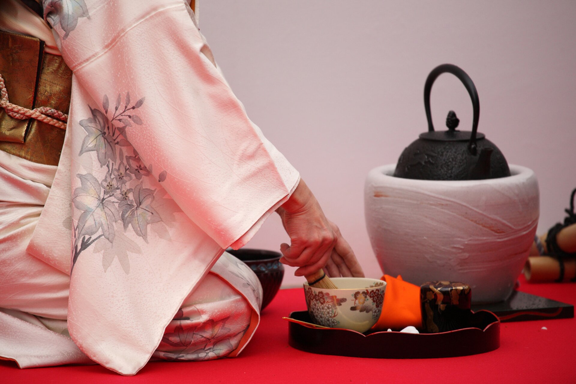 Grafika przedstawia zdjęcie kobiety ubranej w tradycyjny azjatycki strój koloru bladoróżowego z szarymi kwiatami, która przygotowuje herbatę w tradycyjny sposób. Kobieta pokazana jest od pasa w dół i siedzi na kolanach bokiem do kadru. Obok niej znajduje się czarna taca z białą, zdobioną kolorowymi okręgami, porcelanową filiżanką. Za tacą ustawiony jest biały kosz, na którym jest czarny dzbanek z herbatą.