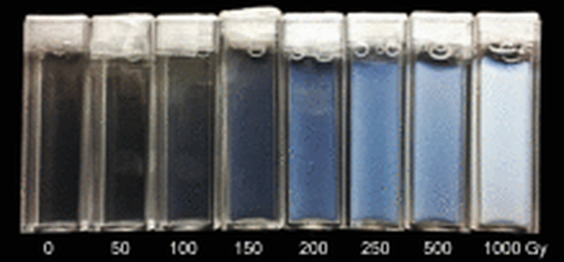 Rys. 7c. przedstawia detektory chemiczne w postaci pojemników z żelem dozymetrycznym na czarnym tle. Pojemniki mają kształt probówek z białym koreczkami u góry. Barwa żelu w środku pojemników zmienia się – od lewej żel jest niemal przezroczysty, następnie przechodzi w barwę niebieską do jasnoniebieskiej. Pod pojemnikami zapis ośmiu dawek promieniowania: 0, 50, 150, 200, 250, 500, 1000 Gy.