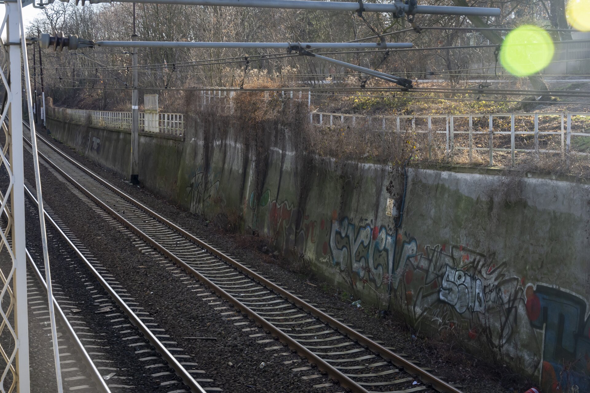  Na zdjęciu widać mur oporowy, który ciągnie się wzdłuż torowiska. Mur został wykonany z żelbetonu. Na jego powierzchni widoczne są graffiti.
