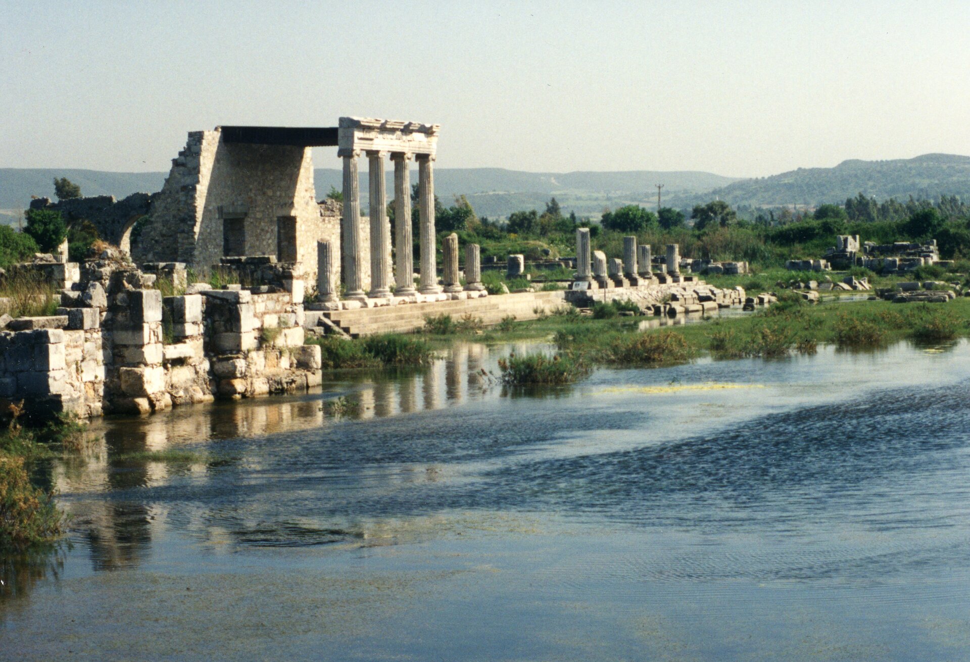 Ilustracja przedstawia ruiny Miletu. Obok zachowanych fragmentów budowli płynie rzeka. W dali widoczne są zarośnięte wzgórza.