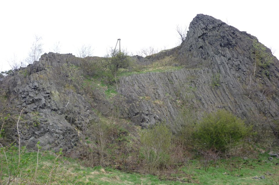 Fotografia przedstawia Czartowską Skałę. Na tle nieba widać niewielkie powulkaniczne wzniesienie, w kształcie małego, lekko skrzywionego stożka.