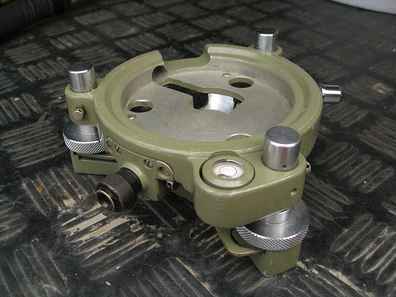 Na zdjęciu znajduje się spodarka z pionownikiem optycznym do geodezyjnych przyrządów pomiarowych. Jest to okrągły element wyposażony w trzy gniazda, w których zamocowane są śruby ustawcze. W jej górnej części znajduje się tuleja z elementem walcowym teodolitu, który blokowany jest śrubą zaciskową. Do dolnej części spodarki przymocowana jest podstawka z jej płytką, zawierającą otwór gwintowany.