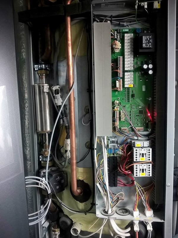 Zdjęcie ukazuje wnętrze sterownika pompy ciepła. Sterownik ten składa się z płytki PCB, czyli elementu elektroniki, modułu sterującego oraz serwisowego wyłącznika głównego. Całość połączona jest cienkimi przewodami.