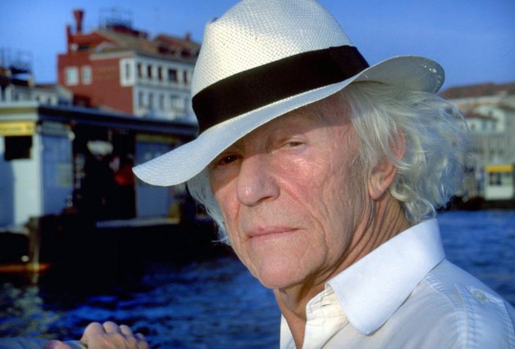 Fotografia przedstawia Romana Opałkę na tle miasta. Opałka to mężczyzna w podeszłym wieku o siwych, falowanych włosach sięgających karku, długim nosie oraz wąskich ustach. Na jego twarzy widoczne są liczne głębokie zmarszczki, jest uchwycony z półprofilu. Ubrany jest w białą koszulę oraz biały kapelusz z czarnym pasem nad rondem. Mężczyzna ze spokojem spogląda w obiektyw aparatu. W tle widoczna jest lekko wzburzona powierzchnia intensywnie niebieskiej wody oraz budynki miejskie. Niebo jest bezchmurne.