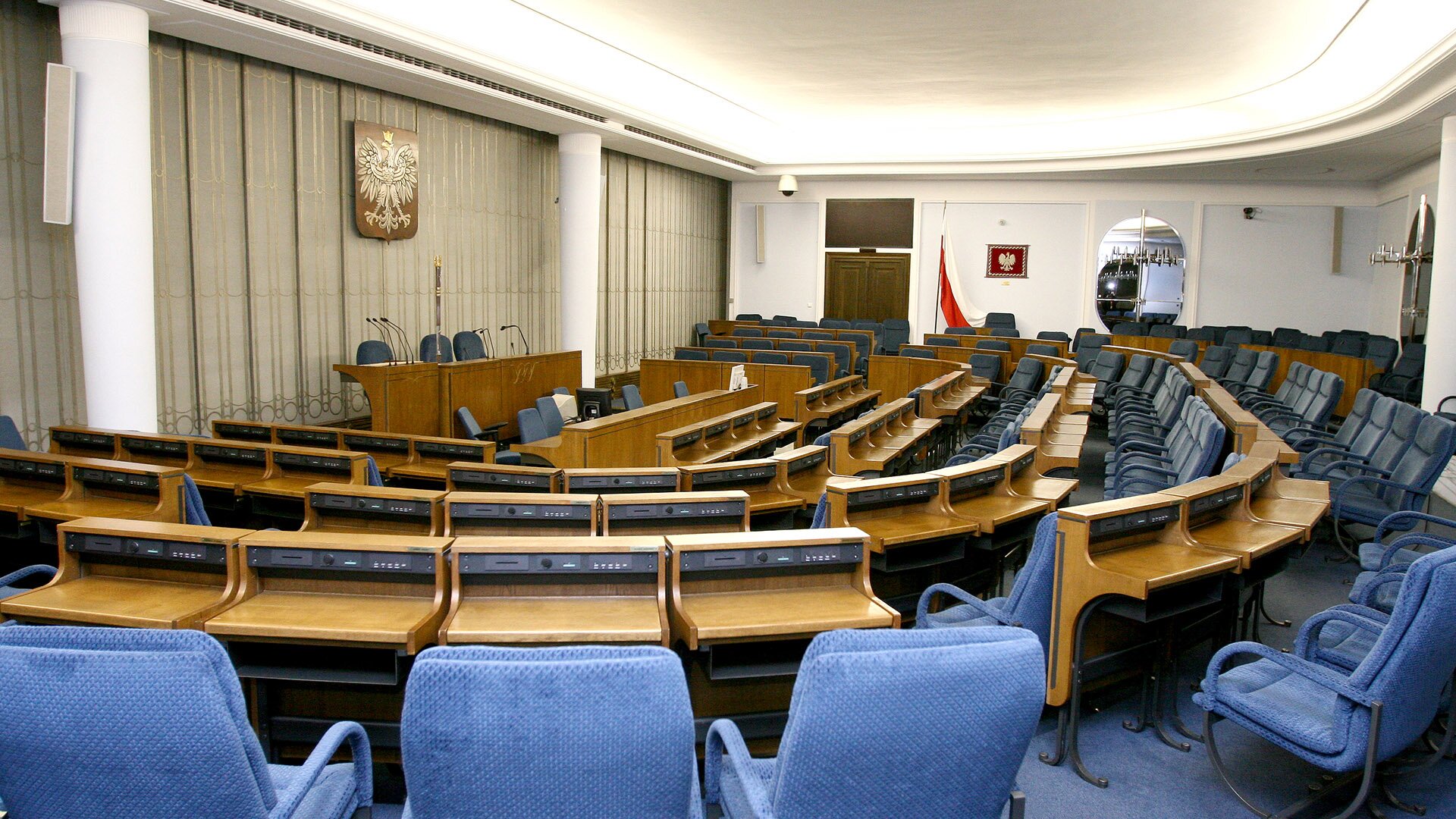 Fotografia przedstawiająca salę posiedzeń Senatu Rzeczypospolitej Polskiej. Po lewej stronie znajduje się stół prezydialny i mównica. Miejsca dla senatorów maja układ amfiteatralny.