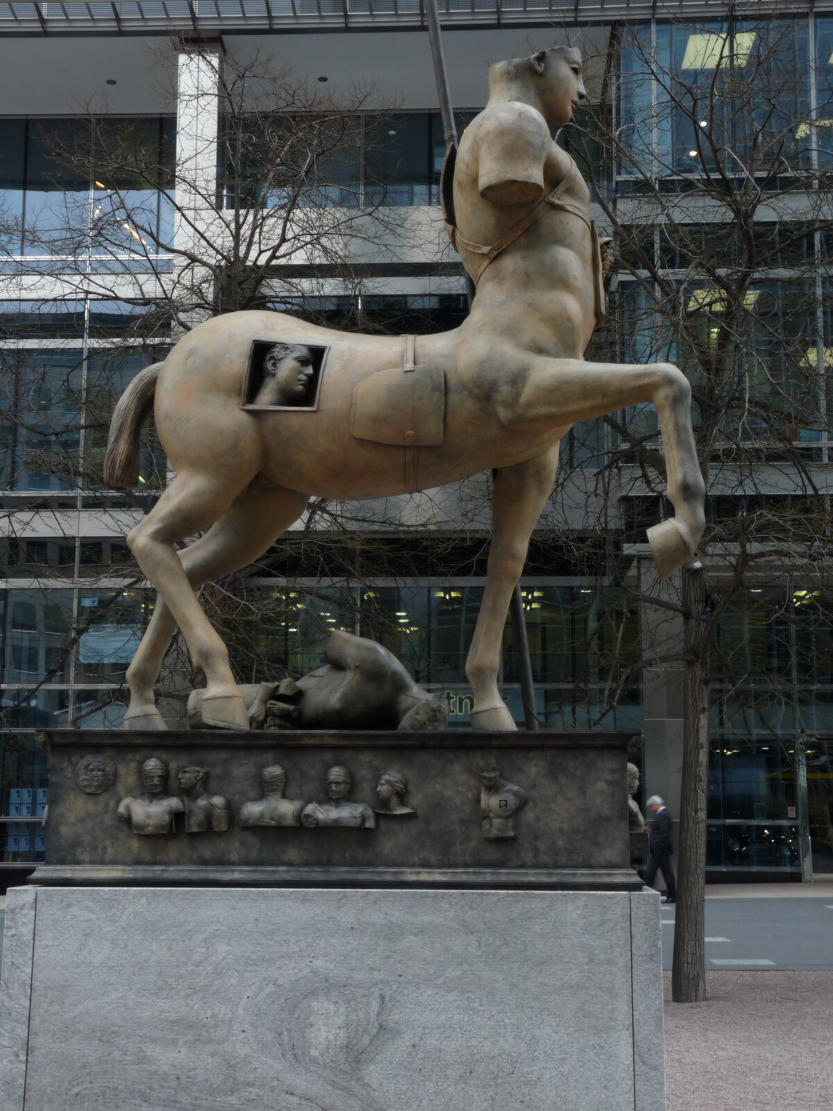Zdjęcie przedstawia rzeźbę centaura stojącego na trzech nogach, czwartą ma uniesioną do góry. Na tułowiu centaura znajduje się ludzka głowa. Centaur nie posiada ręki oraz części głowy. W tle znajdują się drzewa bez liści oraz budynek z dużą ilością okien.