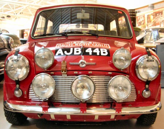 Fotografia przedstawia czerwony samochód MINI, który posiada pełno zamontowanych świateł z przodu. Można mieć wrażenie, że jest to stare auto, które odrestaurowano.