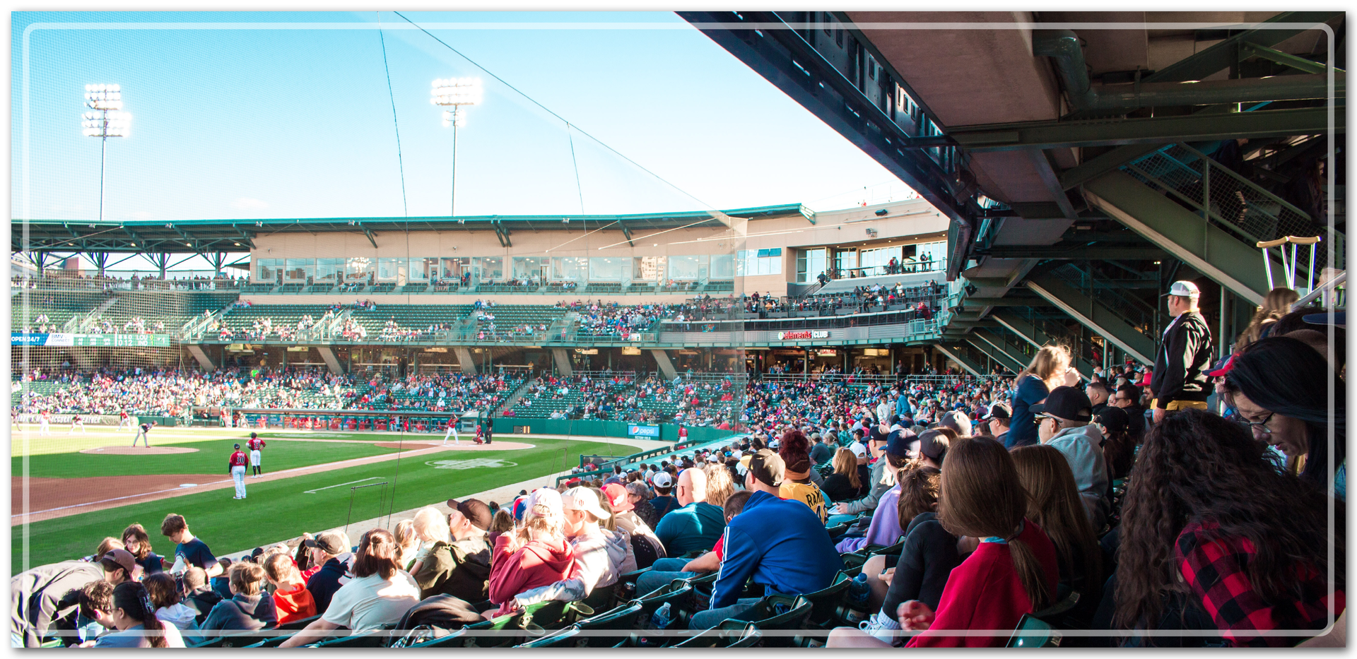 Ilustracja przedstawia tłum ludzi na trybunach przy boisku do Baseball. Zawodnicy w czerwonych koszulkach i białych spodniach stoją w wyznaczonych miejscach na boisku. 