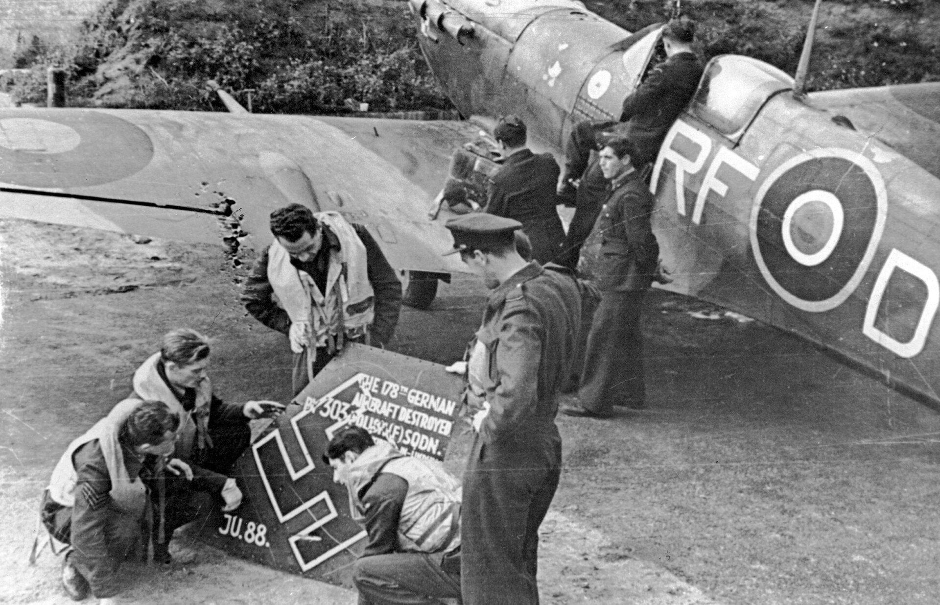 Zdjęcie przedstawia kilku mężczyzn w wojskowych mundurach lotniczych stojących w pobliżu samolotu Spitfire. Kilku z nich ogląda fragment niemieckiego junkersa . Przyglądają się swastyce i napisom, które się tam znajdują.