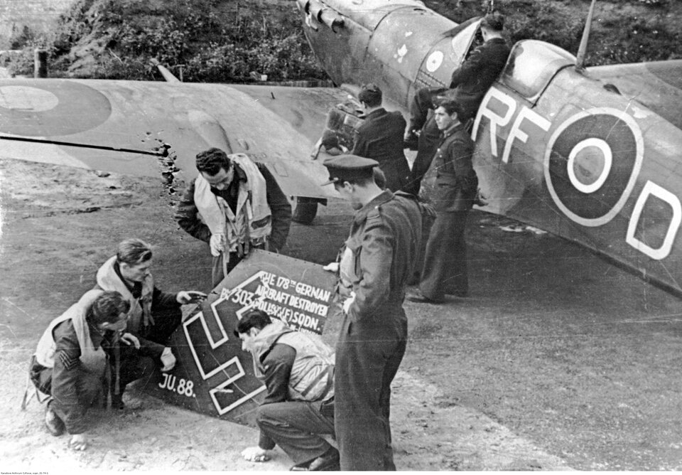 Zdjęcie przedstawia kilku mężczyzn w wojskowych mundurach lotniczych stojących w pobliżu samolotu Spitfire. Kilku z nich ogląda fragment niemieckiego junkersa . Przyglądają się swastyce i napisom, które się tam znajdują.