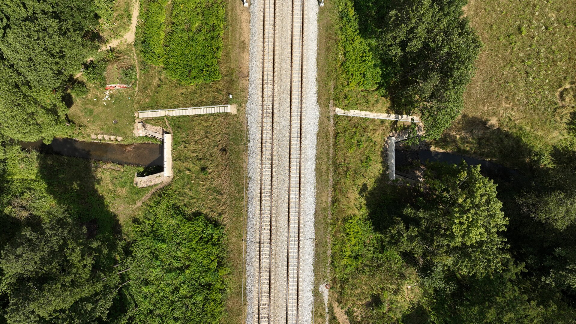 Ilustracja przedstawia przepust widoczny z lotu ptaka. Jest on niskim i wąskim tunelem wbudowanym w nasyp kolejowy, przez który przepływa strumień. Po prawej stronie zdjęcia widać schody prowadzące na szczyt nasypu. Na nasypie widoczne są dwa tory.