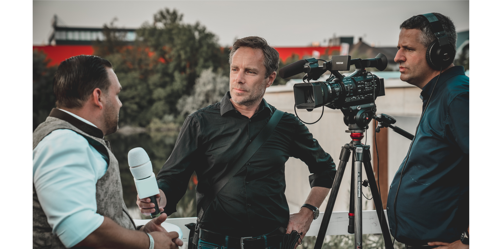 Zdjęcie przedstawia trzech mężczyzn. Po prawej stronie znajduje się operator z kamerą. W środku widoczny jest dziennikarz z mikrofonem w ręku. Dziennikarz przeprowadza wywiad z mężczyzną, który stoi po lewej stronie na zdjęciu.