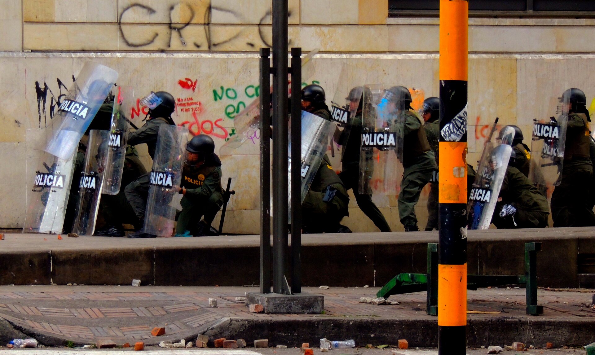 Zdjęcie, które przedstawia interwencję policji. Policjanci ubrani są w kombinezony ochronne, z tkaniny nieprzemakalnej, z odblaskowym napisem policja. Na głowie kask zabezpieczający z osłoną na twarz, pałka służbowa, tarcza ochronna. Policjanci obezwładniają osoby, które uczestniczyły w zamieszkach. Widoczny mur przy którym policja dokonuje zatrzymania. Na murze graffiti. Na ulicy leżące niewielkiej wielkości cegłówki, kamienie.